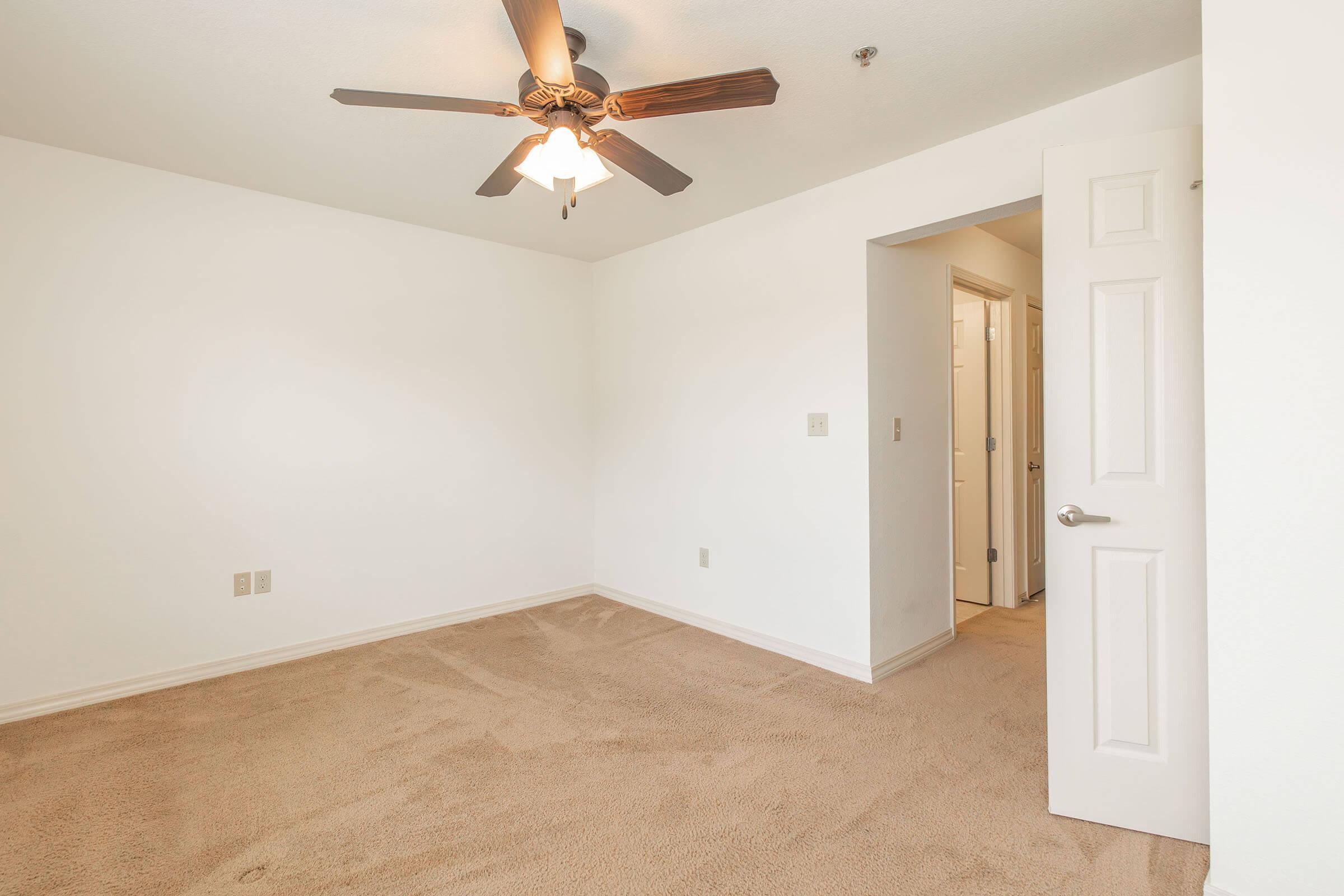 Empty room with beige carpet and white painted walls. A ceiling fan with wooden blades hangs from the ceiling. There is an open door leading to a hallway at the back, revealing additional doors and a light-colored wall. Natural light illuminates the space.