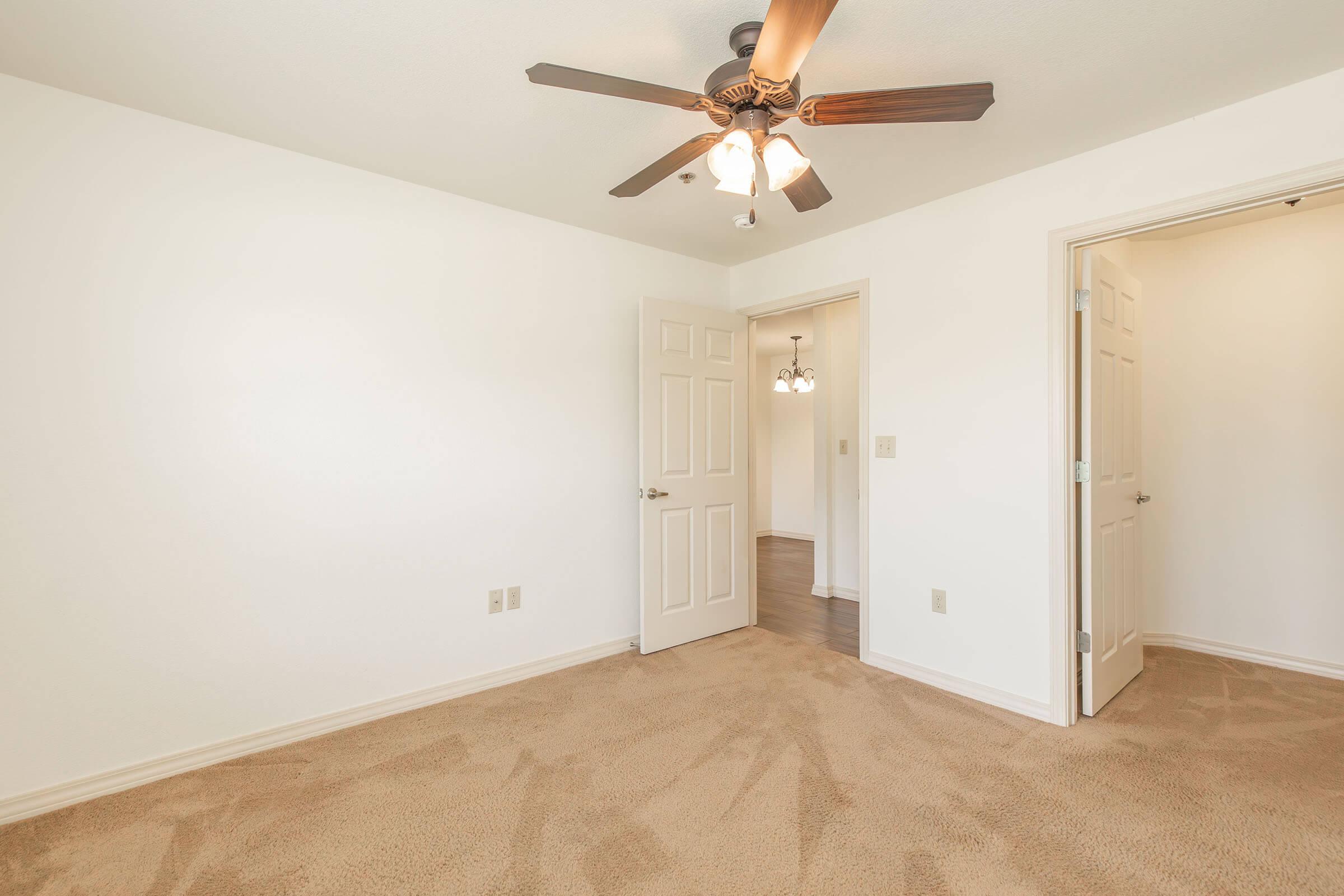 A spacious, empty room with light beige carpet and white walls. A ceiling fan with lights is mounted in the center, and two doorways lead to adjacent rooms. Natural light fills the space, creating a bright, airy atmosphere.