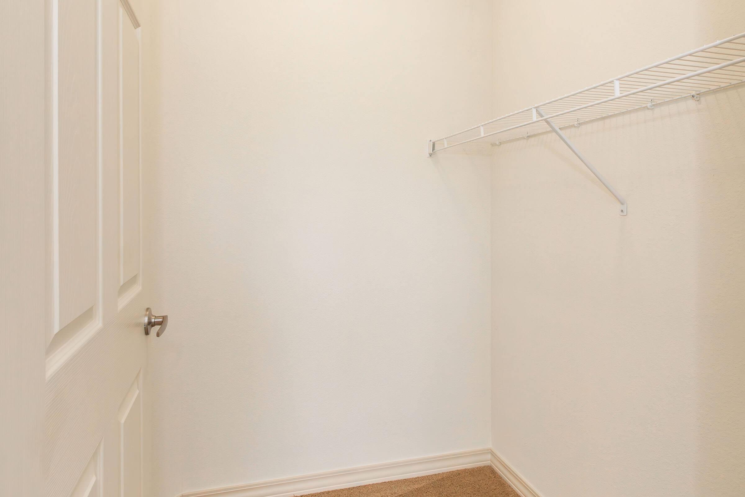 A small, empty closet with a light-colored wall, a single door, and a metal wire shelf mounted on one side. The floor is carpeted, and there is no visible furniture or items inside the closet, creating a minimalist and spacious appearance.