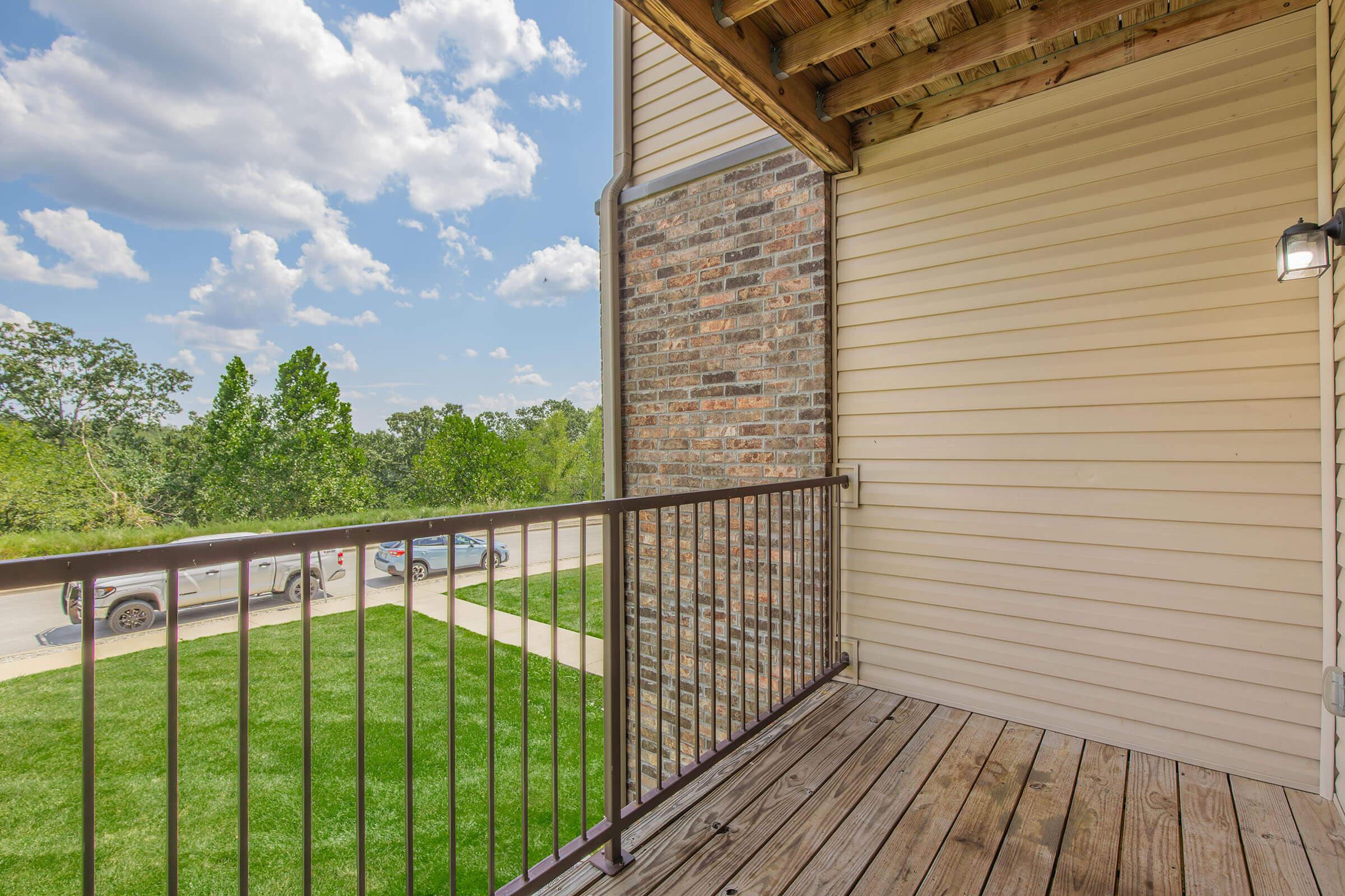 A balcony view featuring wooden flooring, a metal railing, and a brick wall. The setting overlooks a green lawn and trees, with a cloudy blue sky above. Nearby, parked vehicles are visible along a pathway.
