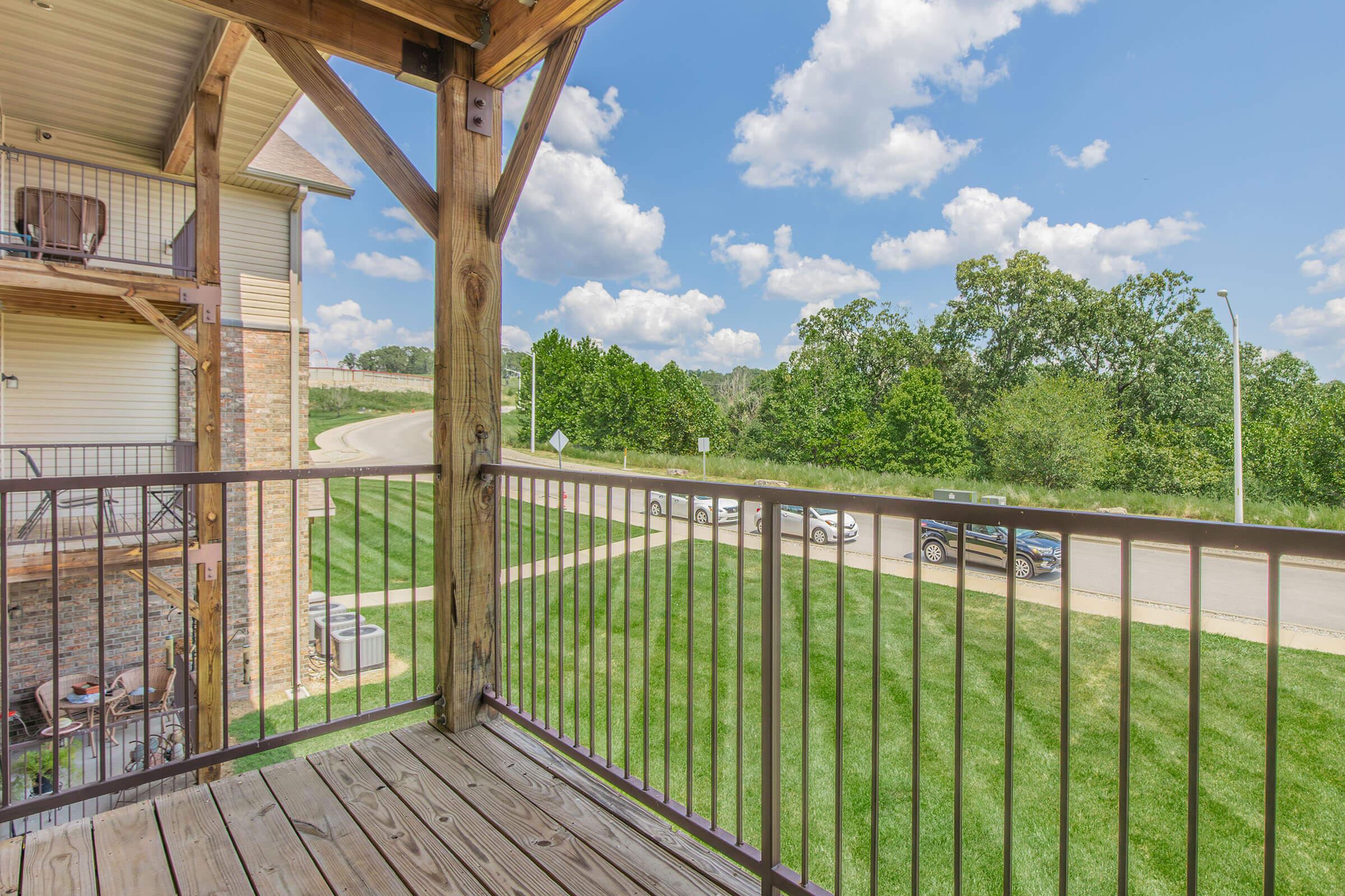 View from a balcony showing wooden railing and grassy area below. In the background, trees and a road are visible under a blue sky with scattered clouds. Several parked cars are seen along the side of the road. The scene conveys a peaceful suburban atmosphere.