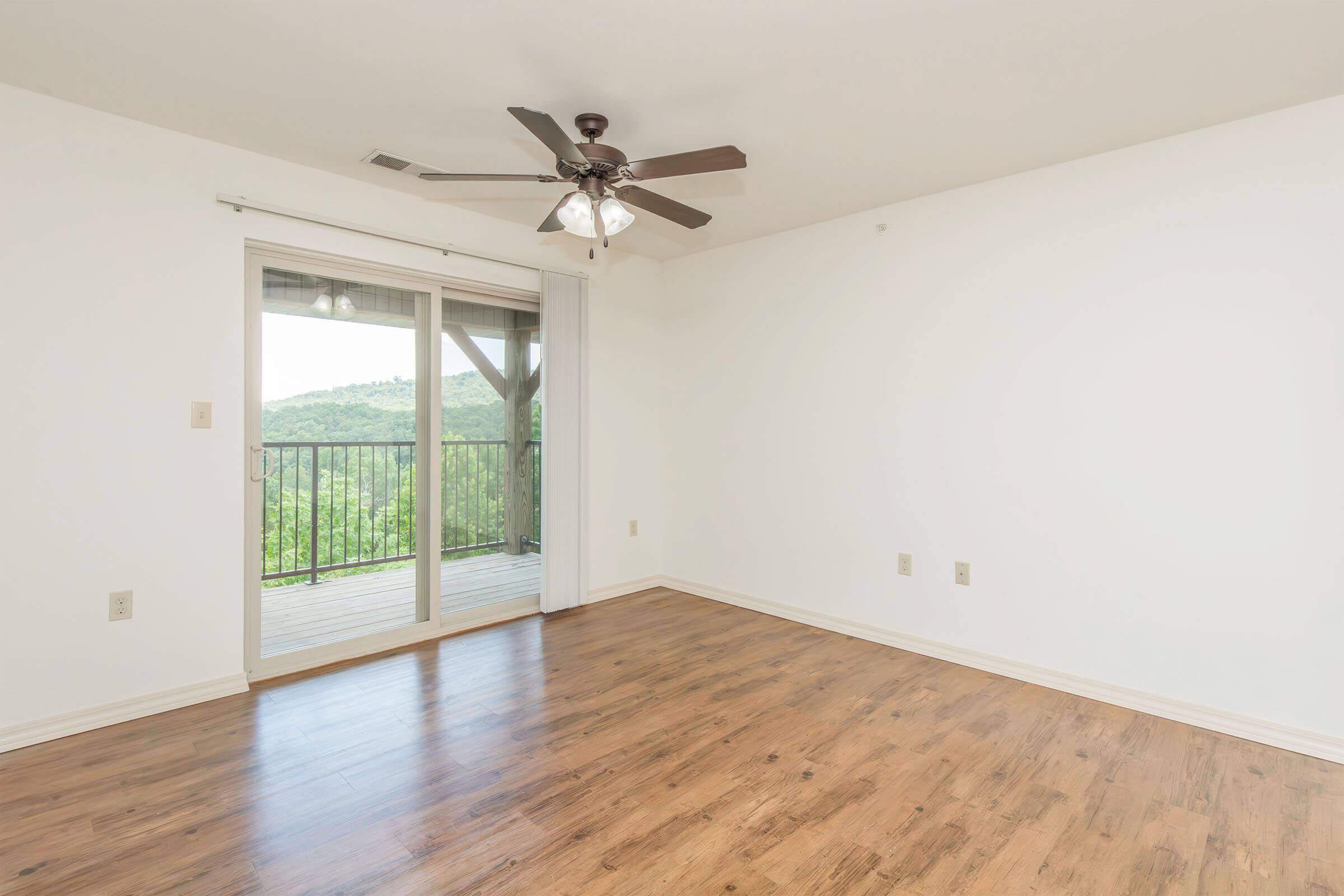 Empty room with light-colored walls and a wood floor. A ceiling fan is visible in the center, and there is a sliding glass door that leads to a balcony with a view of green hills. The space is bright and spacious, with no furniture present.