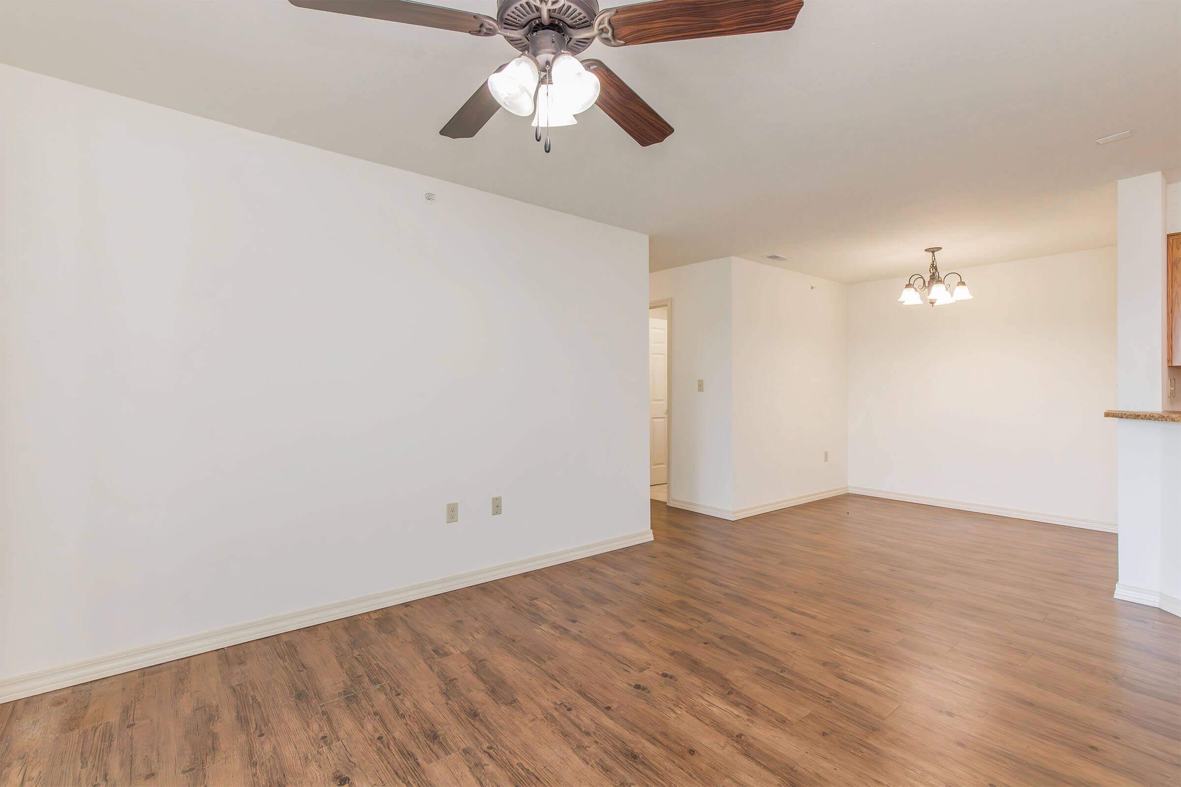 Empty living space featuring light-colored walls, a wooden floor, a ceiling fan with light fixtures, and a hall leading to another room. There is a visible dining area with hanging lights in the background.