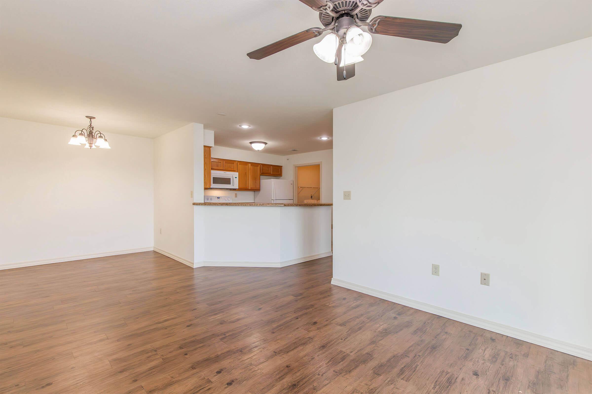 Spacious living area with hardwood flooring, a ceiling fan with light fixture, and an open layout leading to a kitchen area. The kitchen features light wood cabinets and modern appliances, complemented by warm lighting. The walls are painted white, enhancing the bright atmosphere of the space.