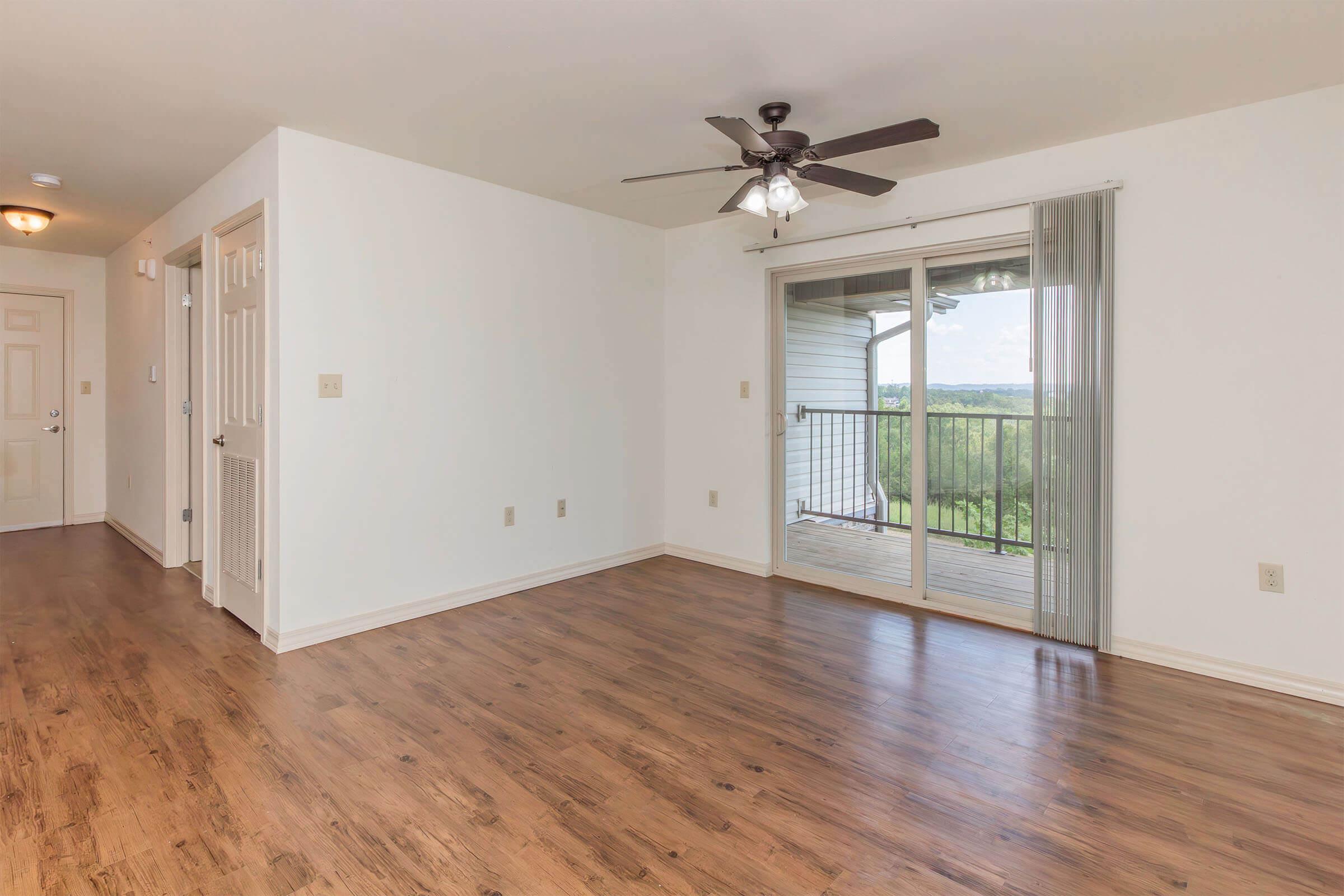 A spacious, empty living room with light-colored walls and a wooden floor. There’s a ceiling fan and a large sliding glass door leading to a balcony with a view. Two doors can be seen on the left side, leading to other rooms or areas. The atmosphere is bright and inviting.