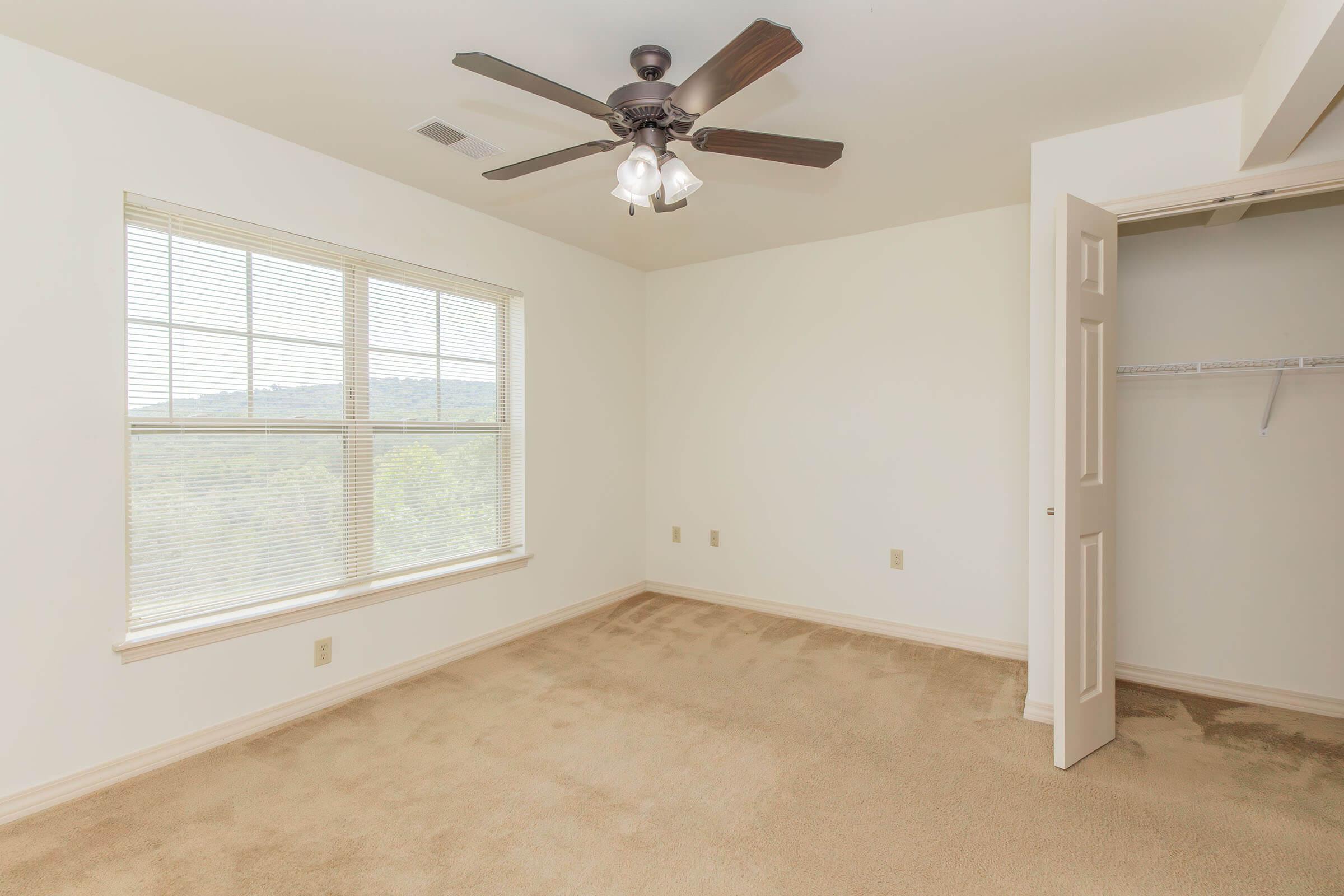 Empty room with light-colored walls and carpet, featuring a ceiling fan with four lights. A large window allows natural light, offering a view of greenery outside. To the right, an open closet with shelving is visible, creating a spacious and airy atmosphere.