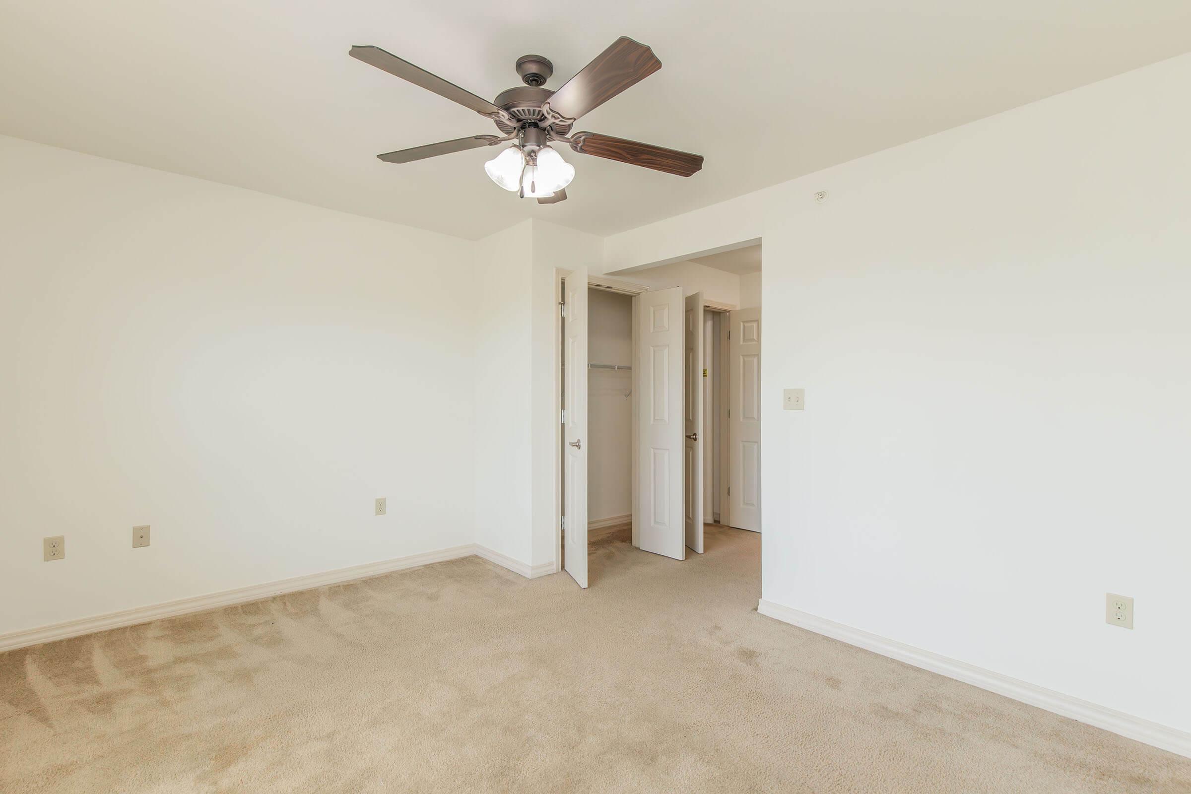 A spacious, empty room with light-colored walls and beige carpeting. A ceiling fan with light fixtures hangs above. There are two open doors leading to adjoining areas, contributing to a bright and airy atmosphere. The room is tidy and uncluttered, ideal for various furnishing options.