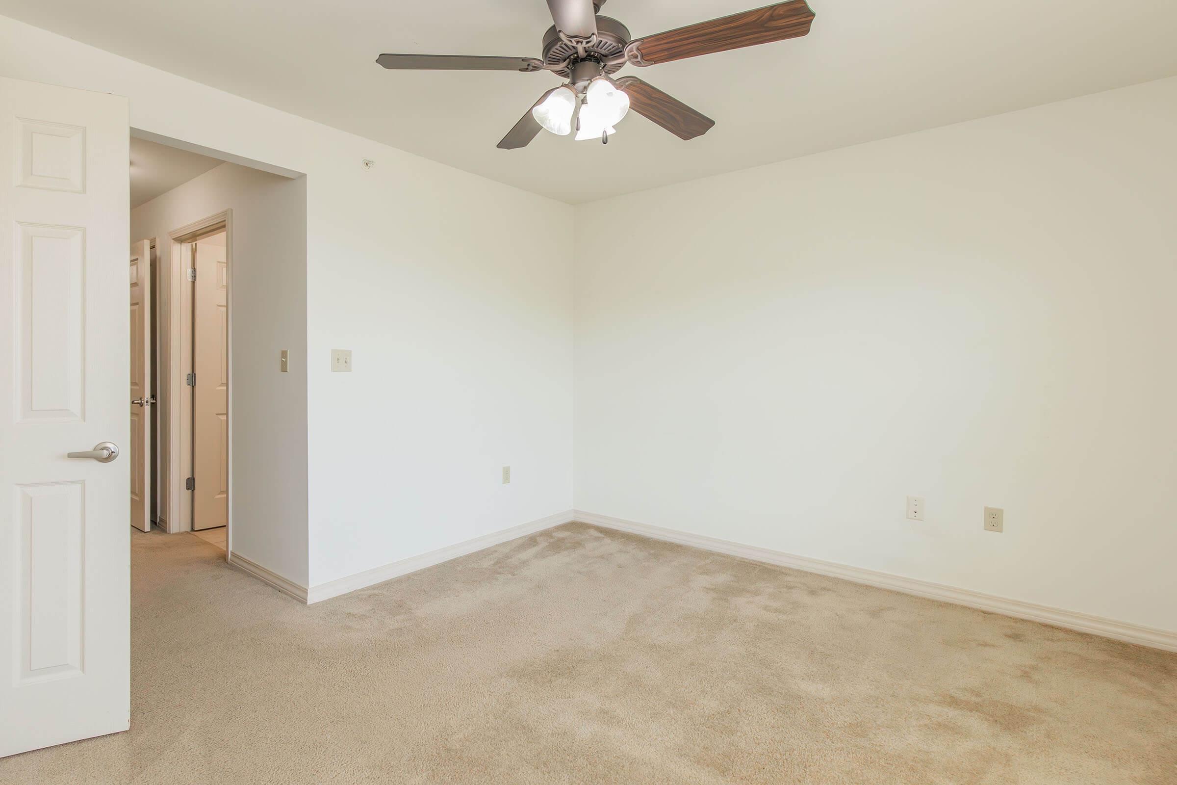 Empty room with light beige carpet, white walls, and a ceiling fan featuring four light bulbs. An open door leads to another area, suggesting potential for use as a bedroom or office space. Natural light may brighten the room, which has minimal furnishings.
