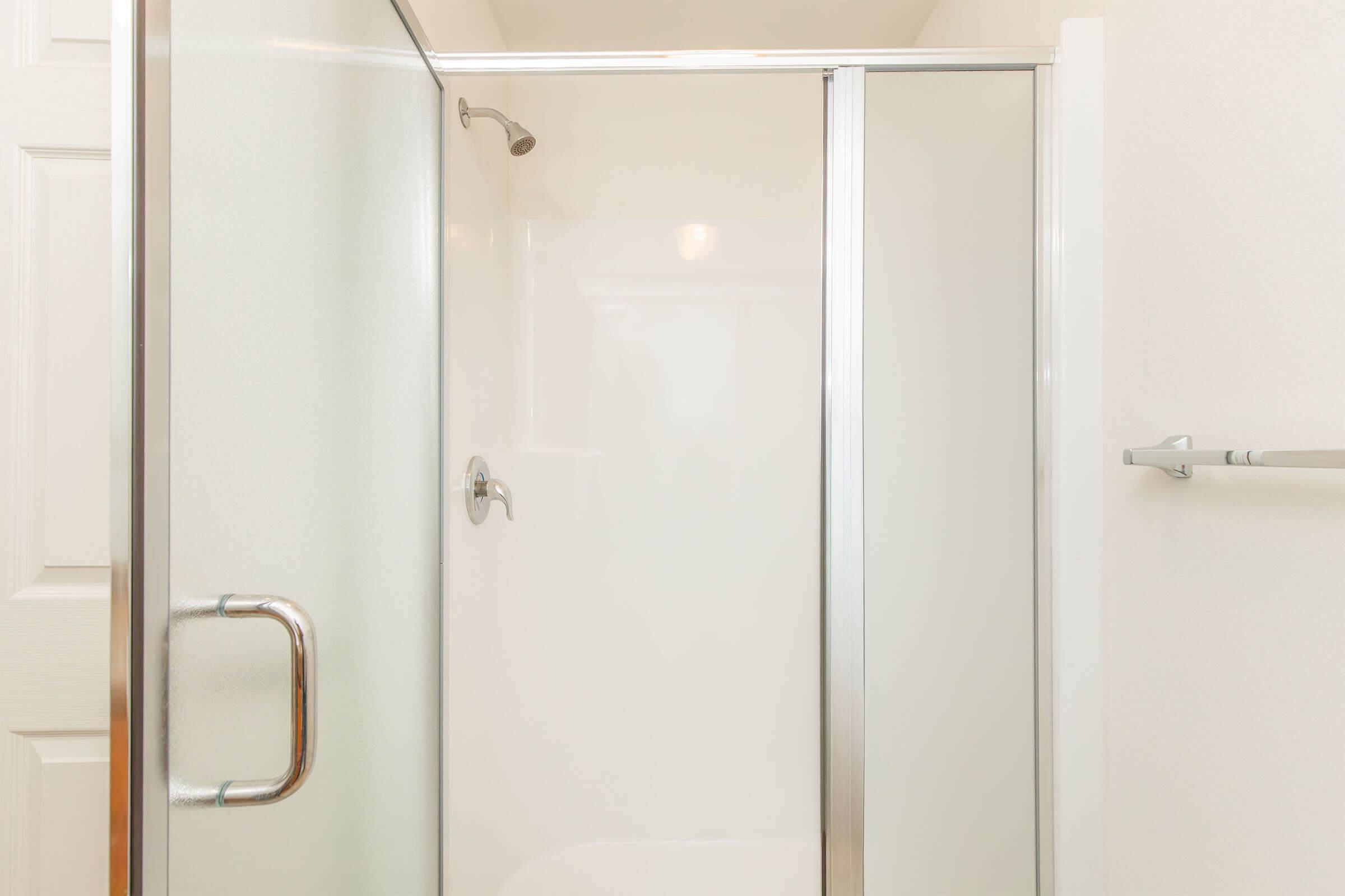 A clean, modern shower stall with clear glass doors and a silver handle. The walls are white, giving a bright and spacious appearance. A towel bar is visible adjacent to the shower.