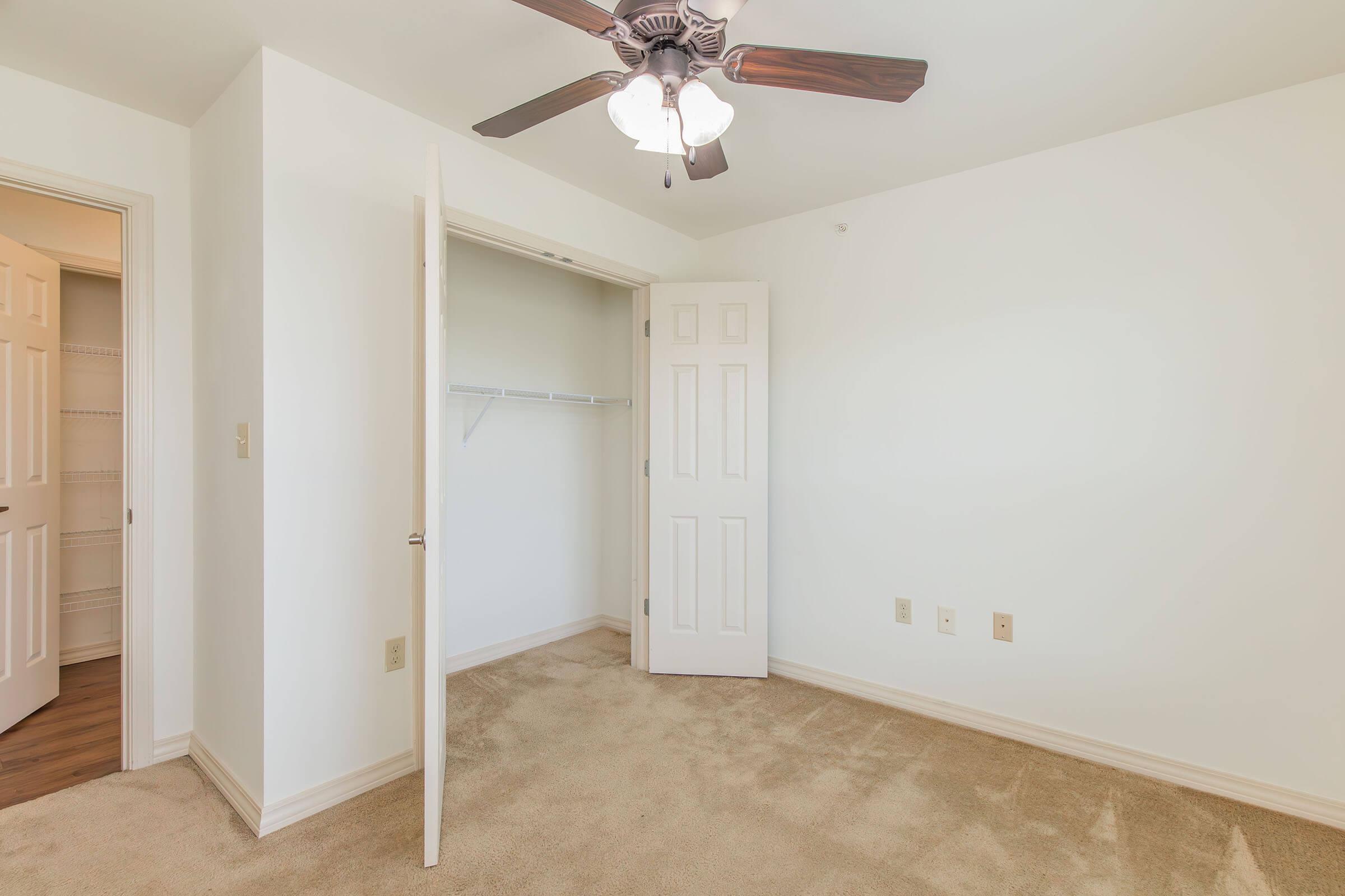 Empty room featuring light-colored walls and beige carpet. A ceiling fan with wooden blades hangs from the ceiling. An open closet with a shelf is visible, along with a partially open door leading to another area. Natural light illuminates the space, creating a bright atmosphere.