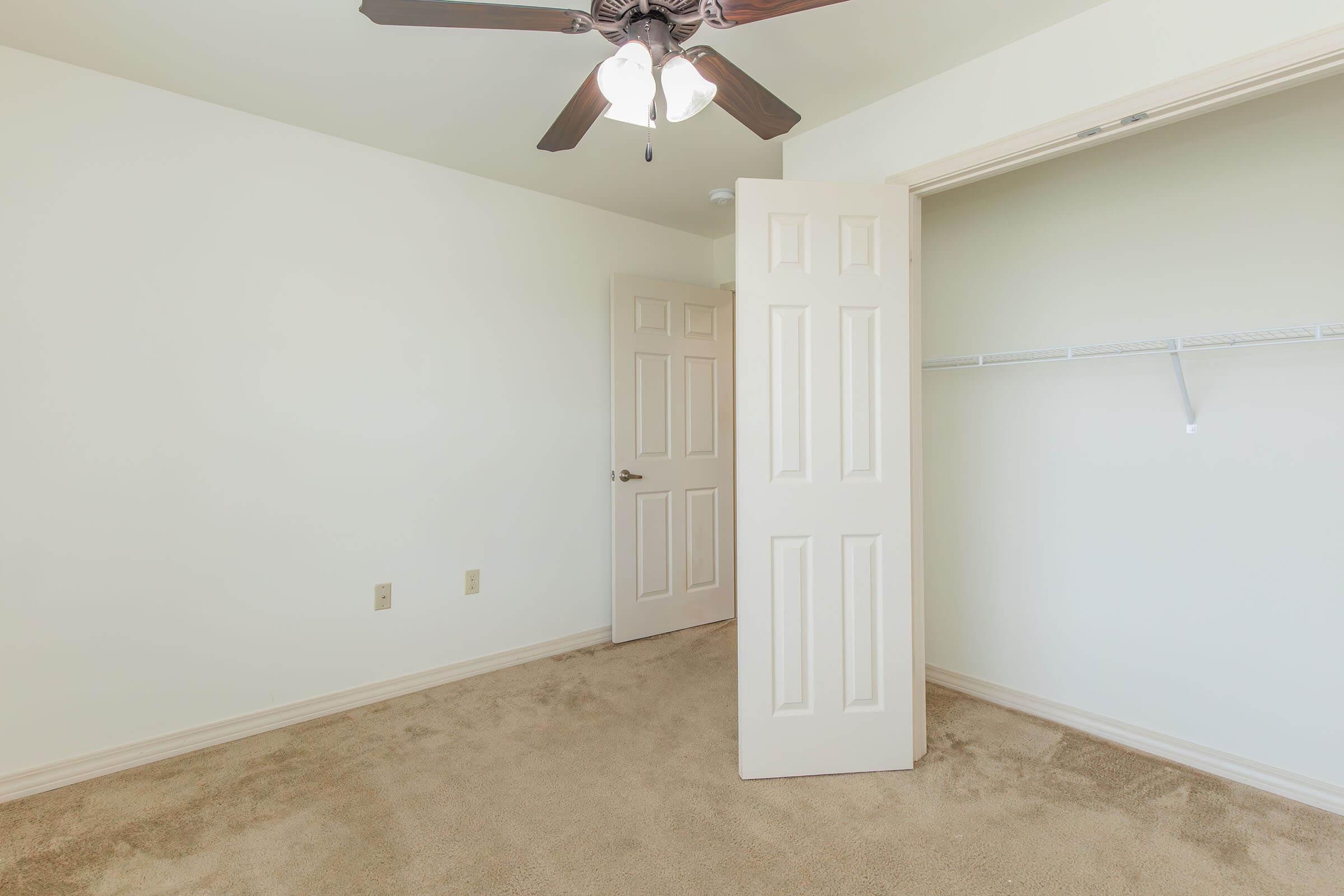 A spacious, empty bedroom with light-colored walls and tan carpet. A ceiling fan with three lights hangs from the ceiling. A door on the left is slightly ajar, revealing a closet with no doors and a shelf. The room has a bright, airy feel, ideal for decorating or personalizing.
