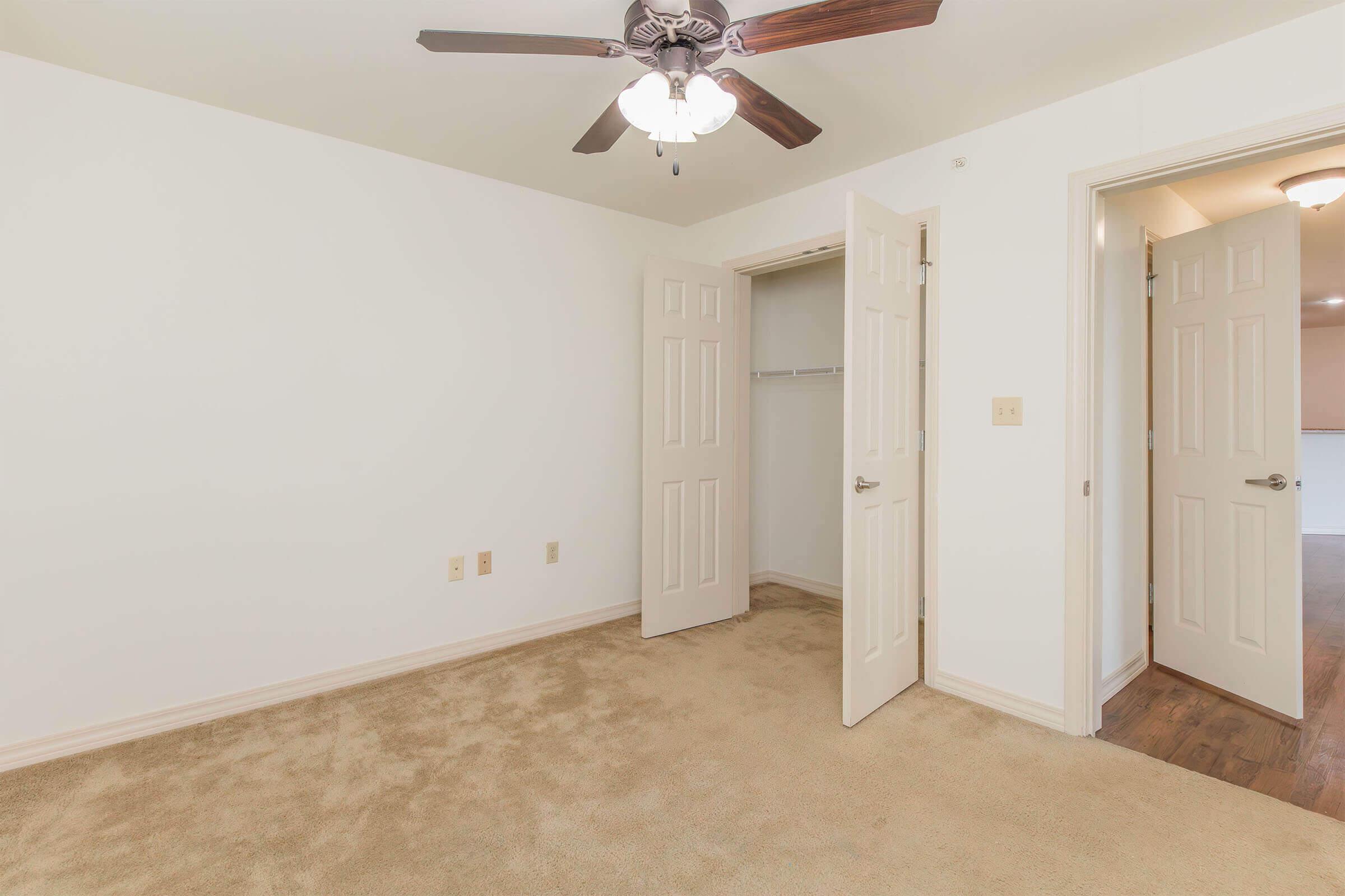 Empty room featuring beige carpet, a ceiling fan with light, and two open doors leading to a closet and another area. The walls are painted white, creating a bright and spacious feel.