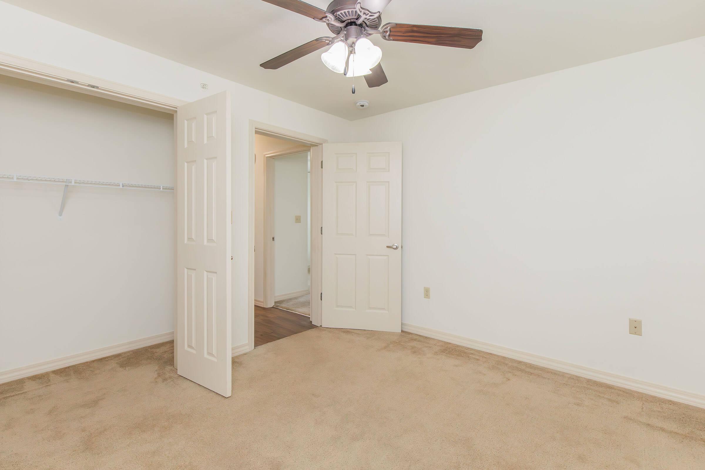 A spacious, empty room with a ceiling fan and cream-colored carpet. Two doors are open, leading to a closet and another room. The walls are painted white, creating a bright and airy atmosphere. No furniture or decorations are present, emphasizing the room's openness and potential for personalization.
