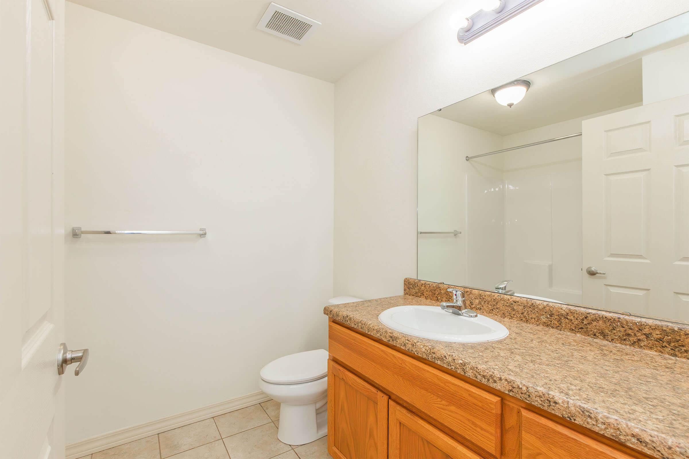 A clean, modern bathroom featuring beige tile flooring, a wooden vanity with a sink, a large mirror above the vanity, and a white toilet. The walls are painted light beige. A frosted glass shower enclosure is visible in the background, and a towel bar is mounted on the wall beside the mirror.