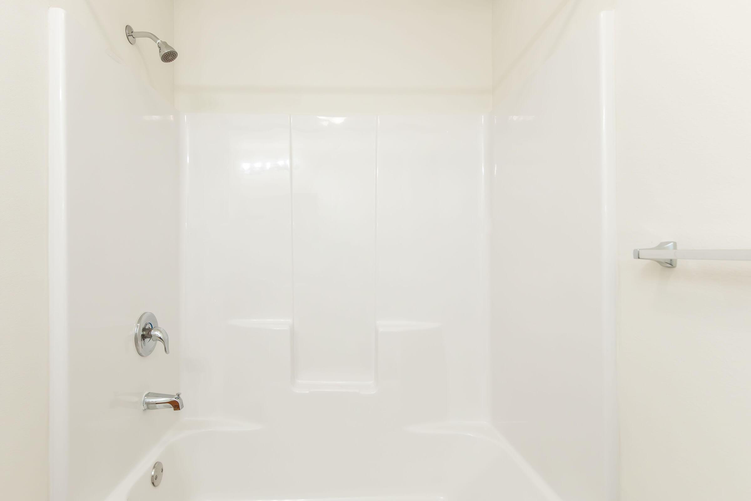 A clean, empty shower stall with a white acrylic tub and walls. The showerhead and faucet are visible on the left side, while a shower grab bar is mounted on the right wall. The surroundings are bright with neutral-colored walls, emphasizing a simple and uncluttered design.