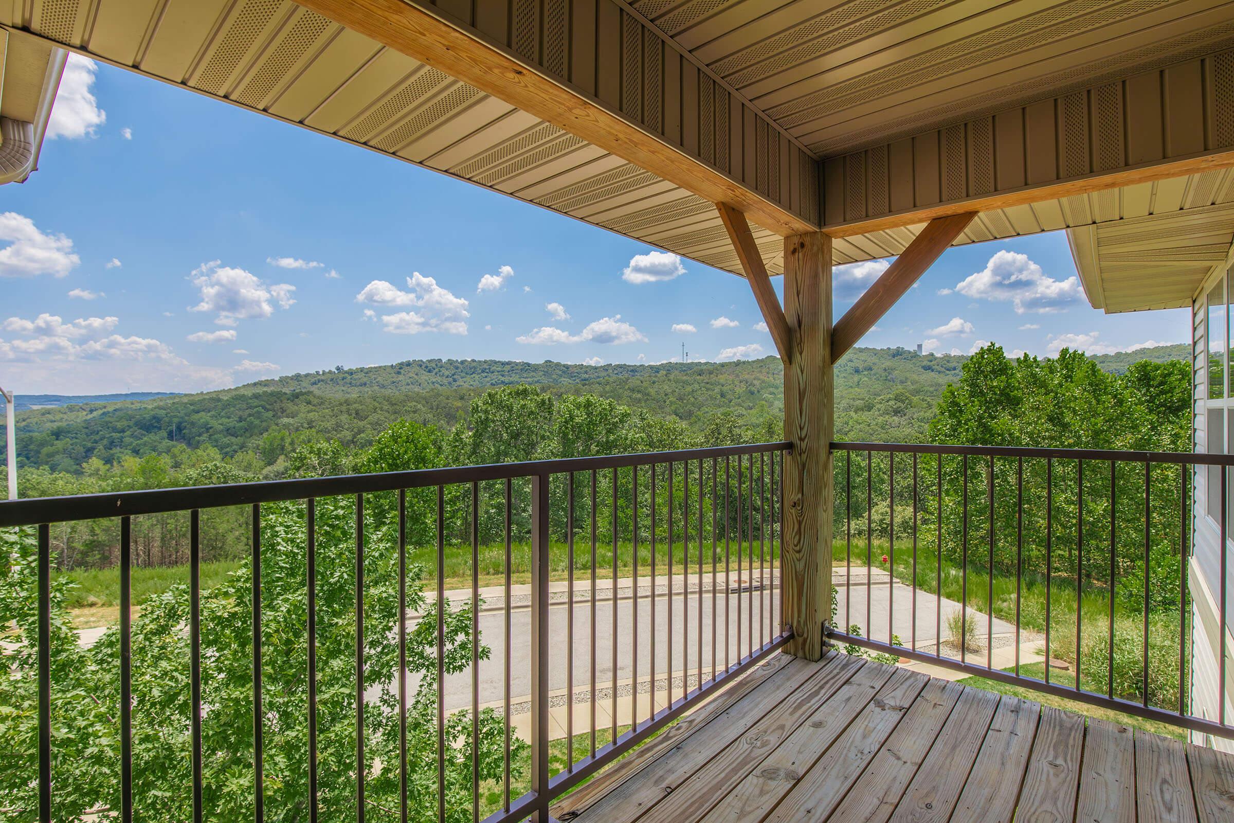 View from a balcony overlooking lush green hills and a clear blue sky with scattered clouds. The wooden railing frames the picturesque landscape, while a small road is visible in the lower part of the image. The scene conveys a peaceful, natural environment.