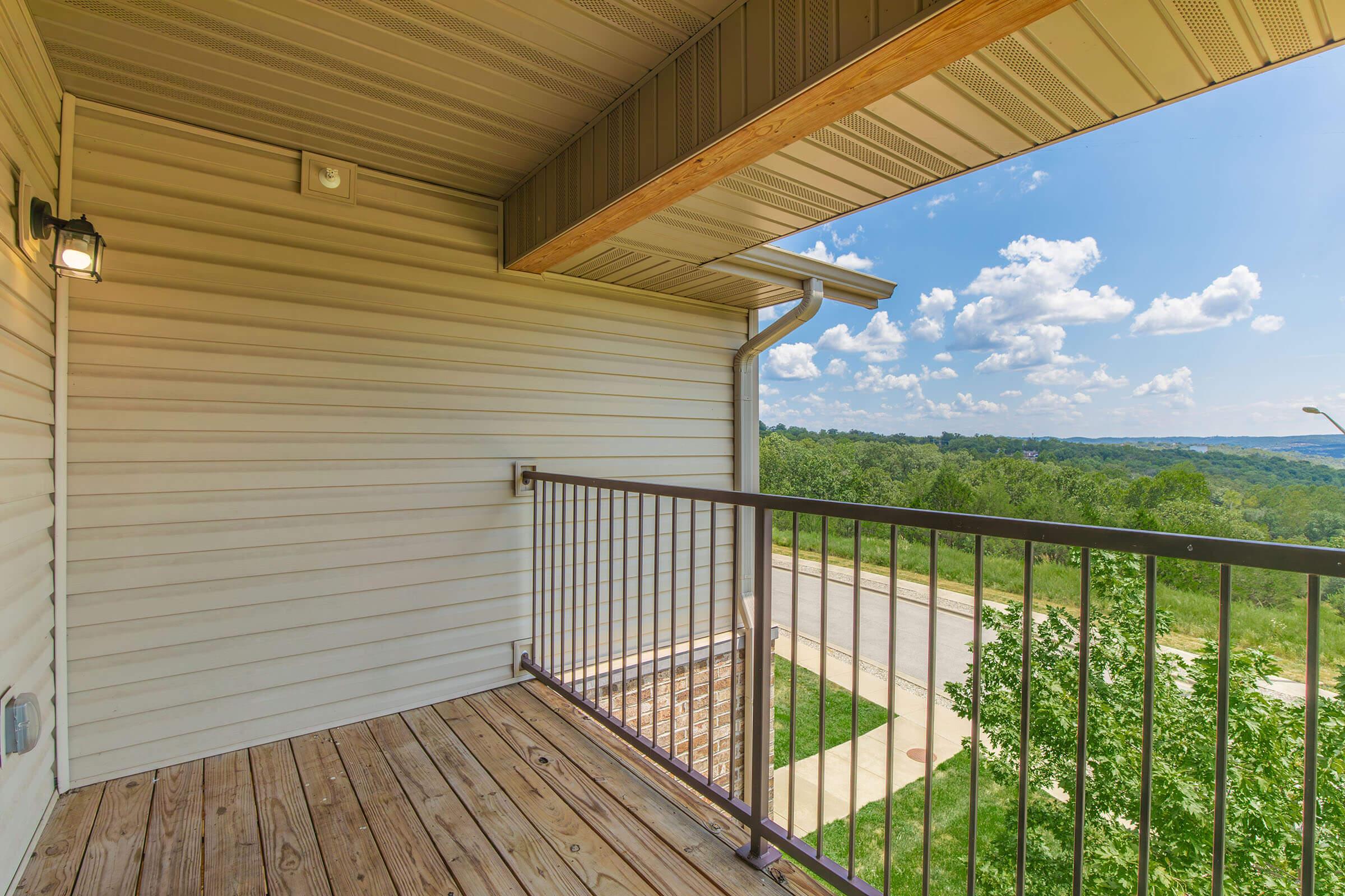 Empty outdoor balcony with wooden flooring, metal railing, and a small light fixture. The view includes trees and a road in the distance under a blue sky with scattered clouds. The balcony is surrounded by beige siding and has an overhang.