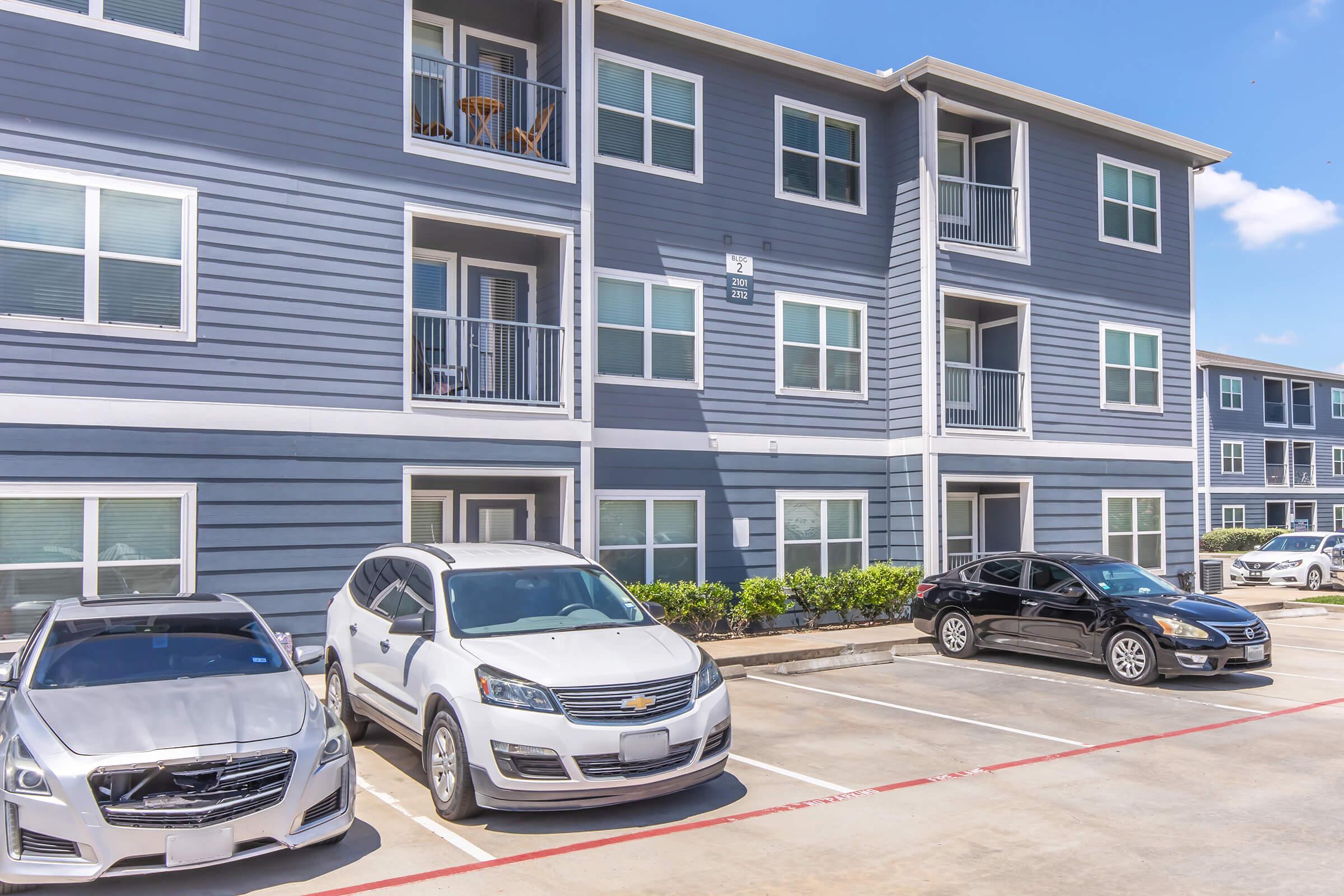 A parking lot in front of a multi-story apartment building. Several vehicles are parked, including a white SUV and a silver sedan. The building has a modern design with multiple balconies and blue-gray siding. There are green shrubs along the parking area under a clear blue sky.