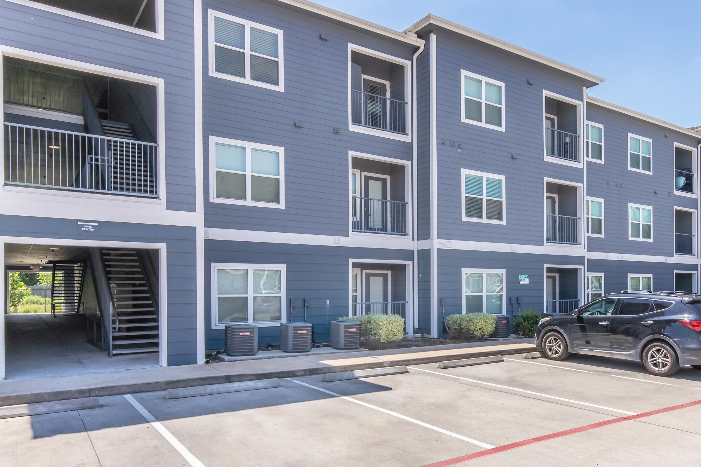A modern apartment building featuring multiple units with blue-gray siding, balconies, and large windows. The ground level includes air conditioning units and a staircase leading to the upper floors. A black SUV is parked in front, with a clear blue sky in the background.
