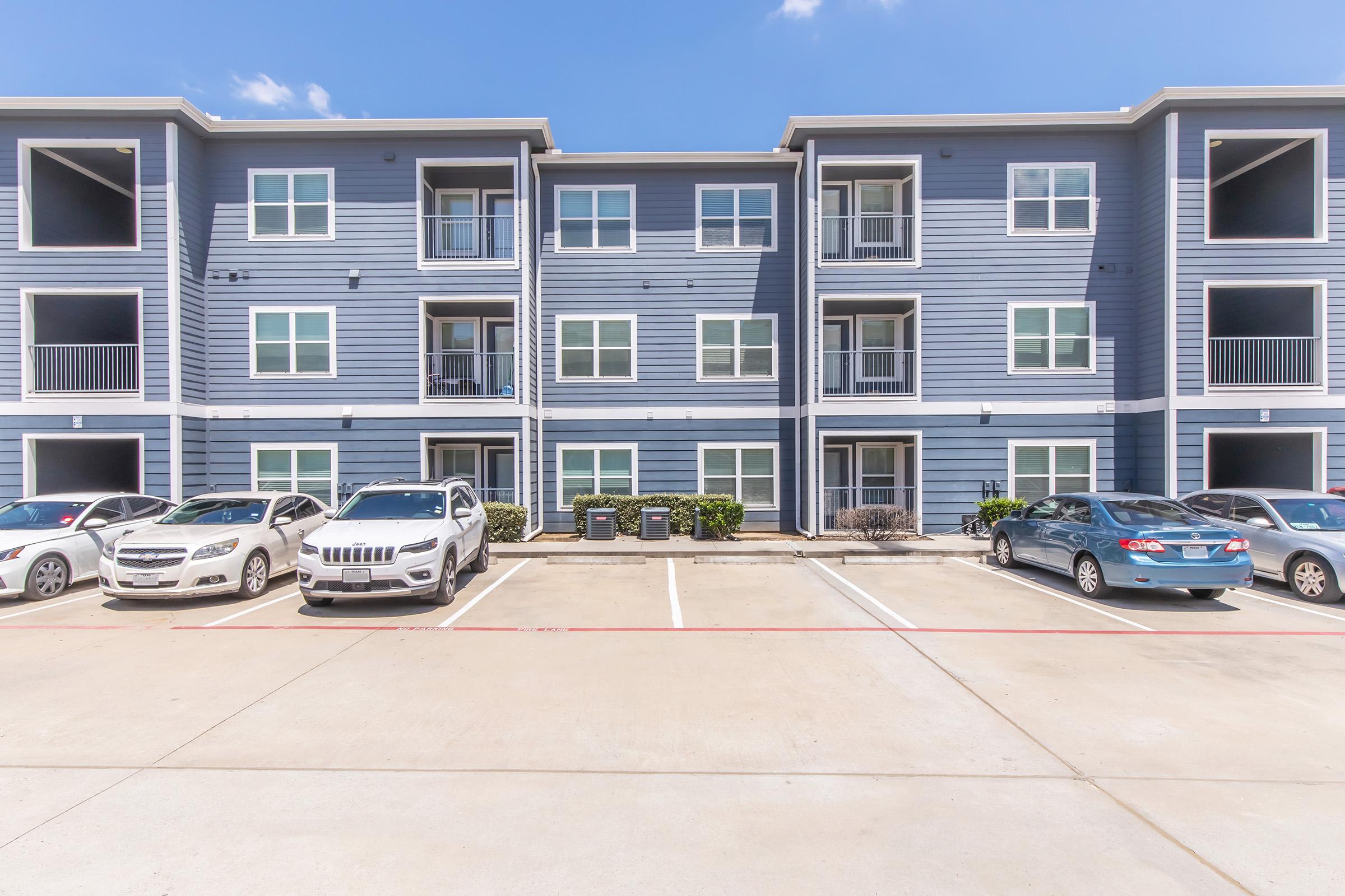 A view of a multi-unit apartment building with blue siding, featuring three levels of balconies. Several parked cars are visible in the front, with a well-maintained parking lot and landscaping. The sky is clear with a few clouds.
