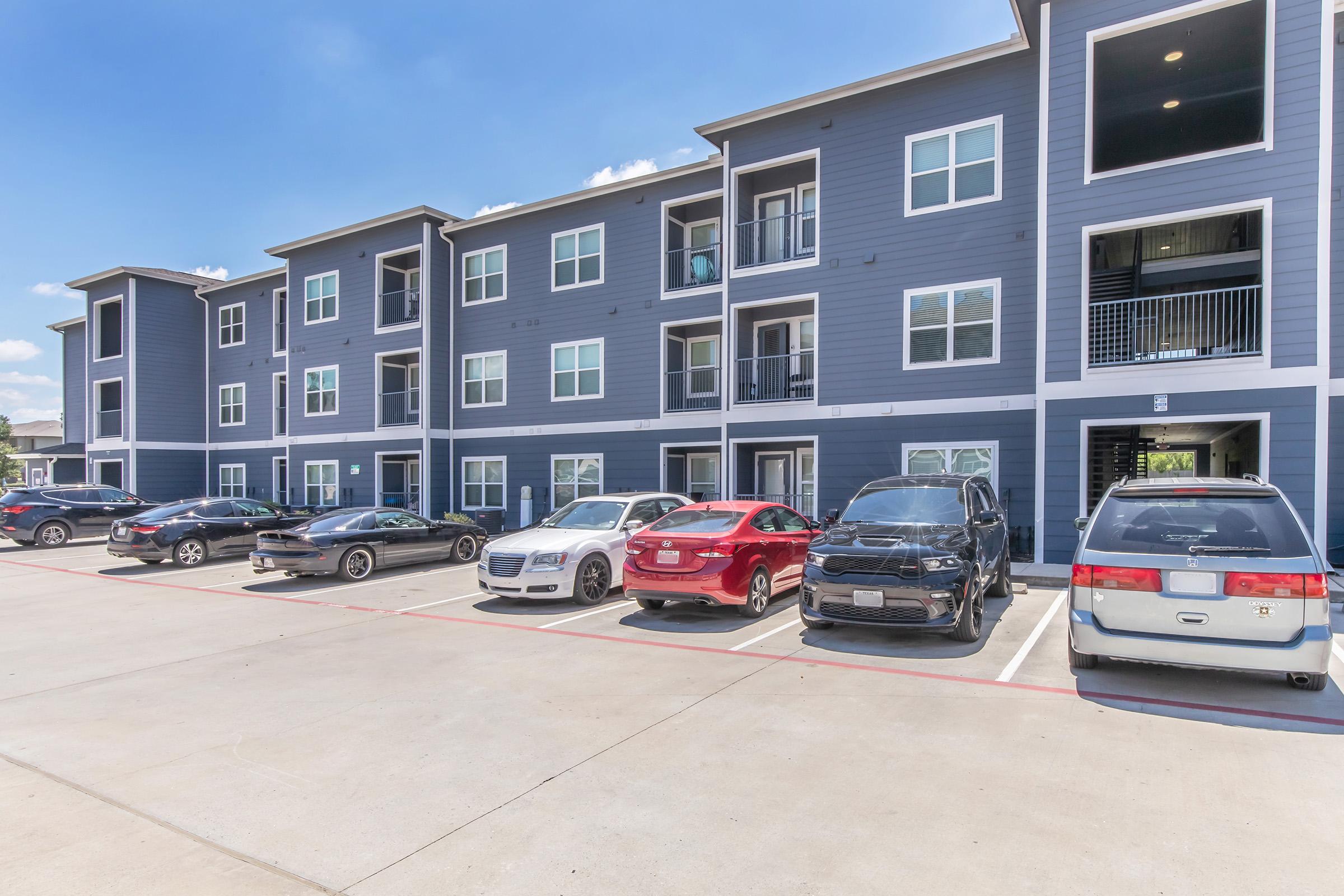 A modern apartment complex featuring multiple two-story buildings with blue exteriors. Several parked cars in front, including a red sedan, a black SUV, and a silver luxury vehicle. The sky is clear and sunny, enhancing the contemporary aesthetic of the property.