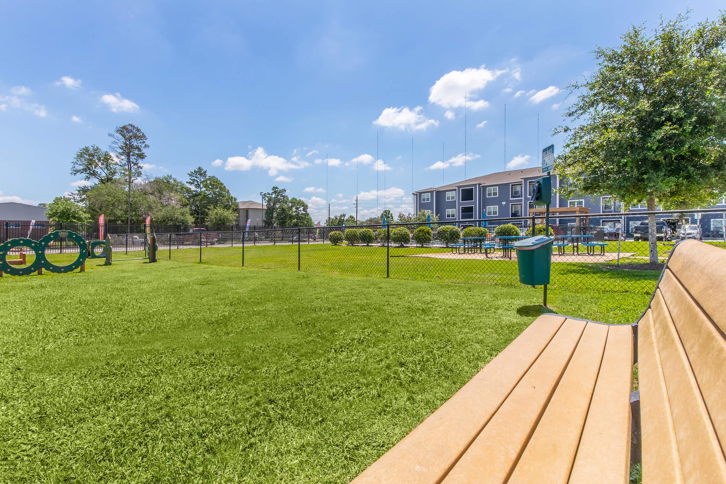 A grassy park area featuring a wooden bench in the foreground. In the background, there are amenities such as a swimming pool and well-maintained landscaping, along with a fenced area and an apartment building. The sky is clear with a few clouds, indicating a sunny day.
