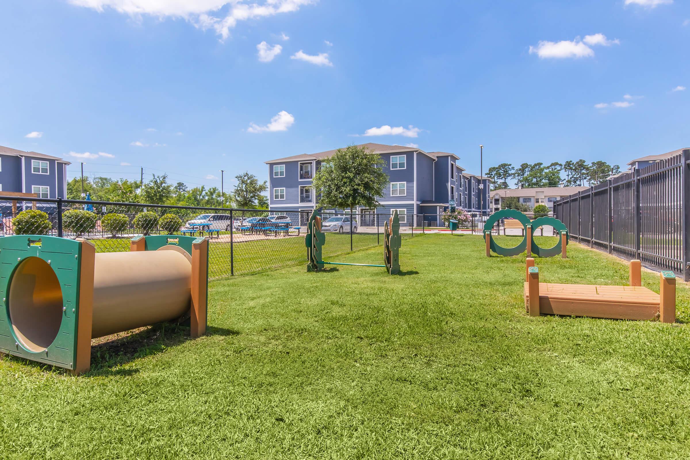 A grassy dog park featuring various agility equipment, including tunnels, ramps, and hoops, set within a fenced area. In the background, there are modern apartment buildings and clear blue skies.