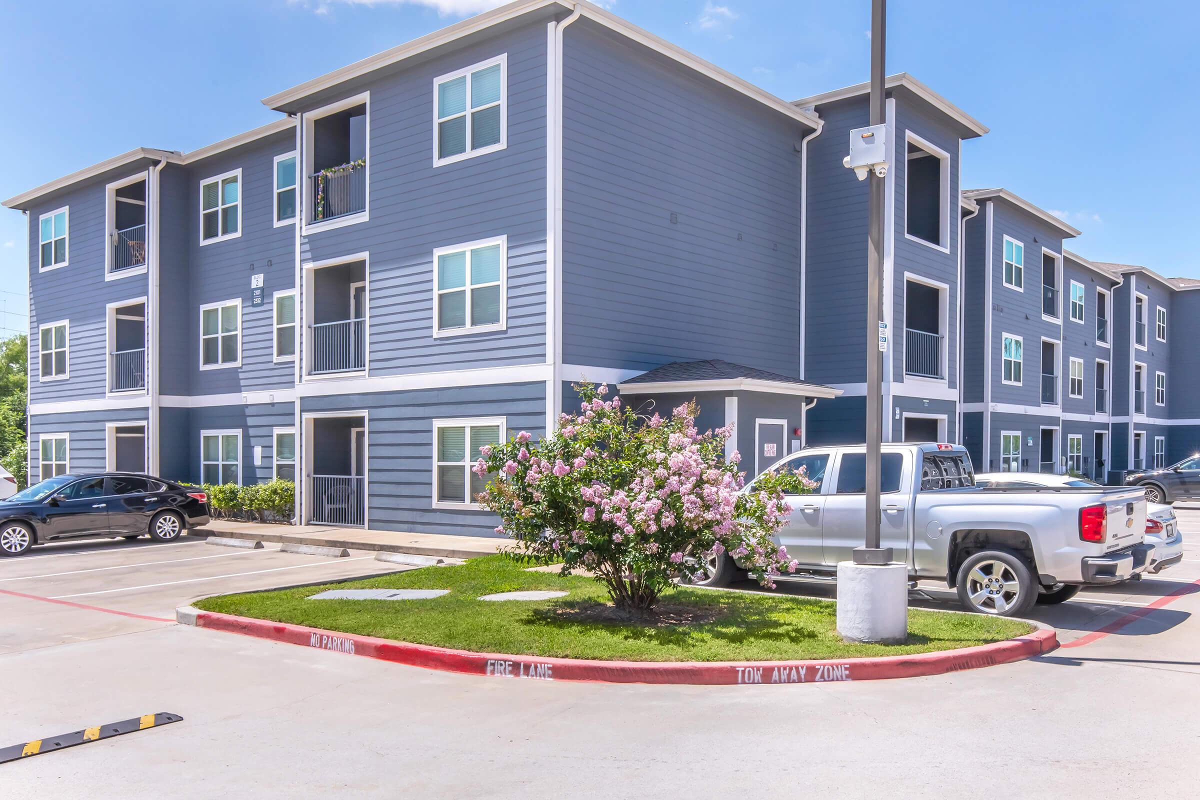 A multi-story residential building with gray siding and white trim. There are flowering plants in front, a parking area with several cars, and bright blue skies above. The building features balconies and a landscaped lawn. A sign indicating "FIRE LANE" is visible near the curb.
