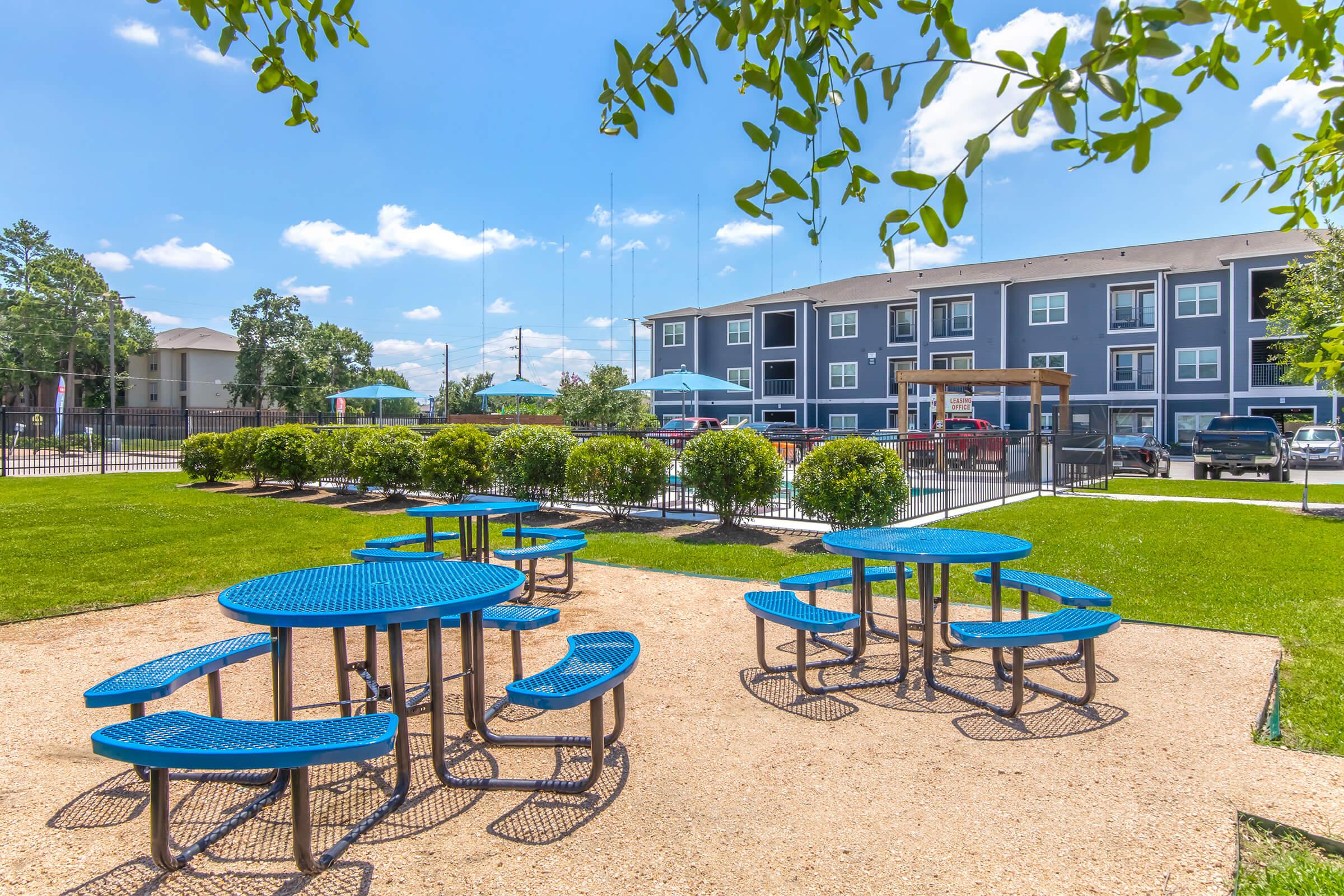 A sunny outdoor area featuring several blue picnic tables surrounded by green grass and shrubs. In the background, there are apartment buildings and a fenced swimming pool area, all under a bright blue sky with scattered clouds.
