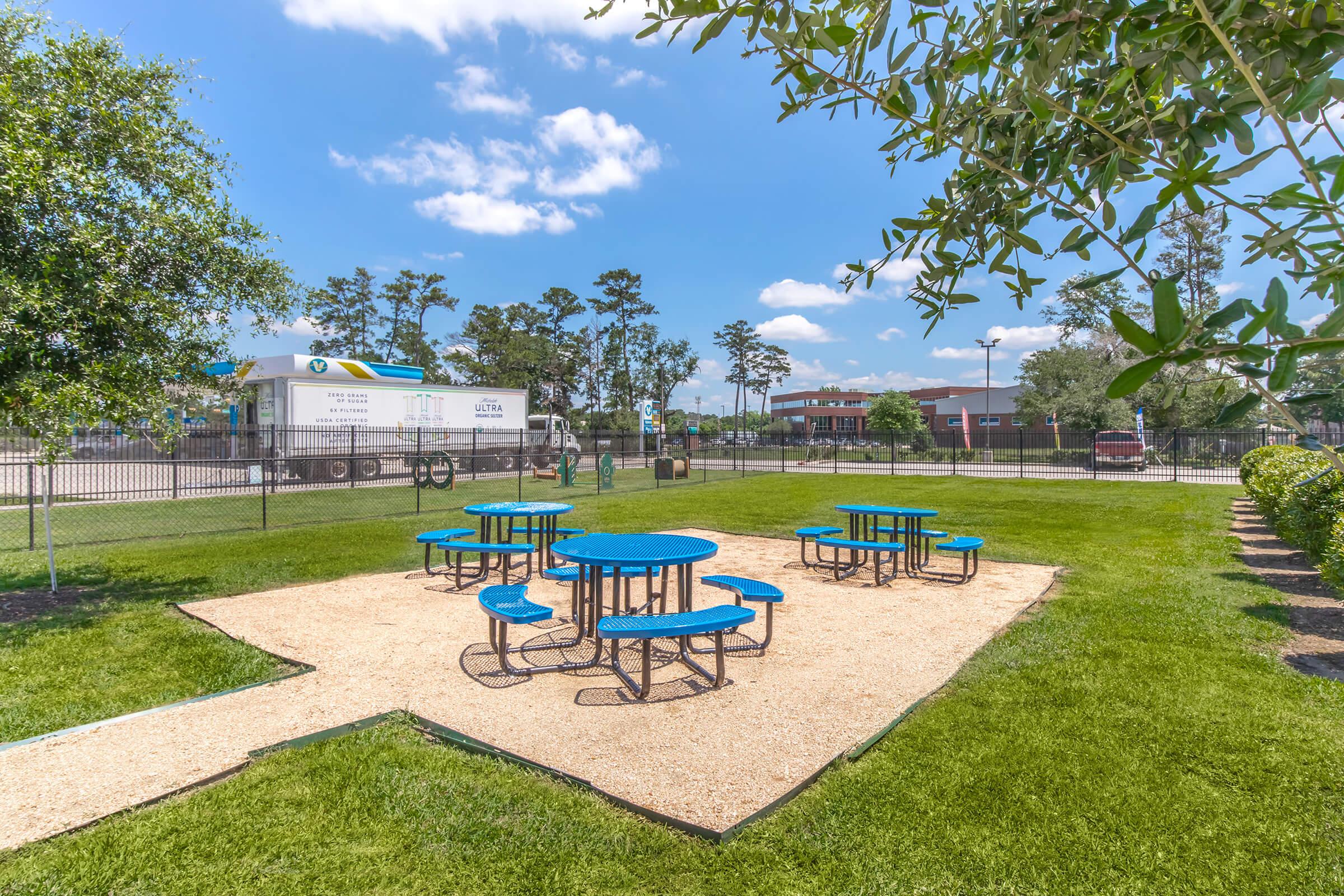 A sunny outdoor picnic area featuring several blue tables and benches on a gravel surface, surrounded by green grass and trees. In the background, a fenced area with a truck visible, indicating a functional outdoor setting suitable for gatherings or breaks. Bright blue sky with a few fluffy clouds.
