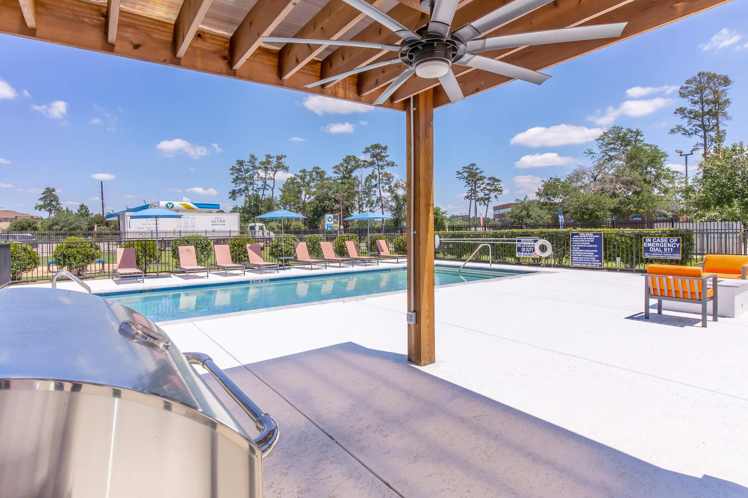 A view of a swimming pool area featuring lounge chairs, a shaded seating area with orange furniture, and a grill underneath a wooden structure. Surrounding greenery and a clear blue sky with a few clouds can be seen in the background.