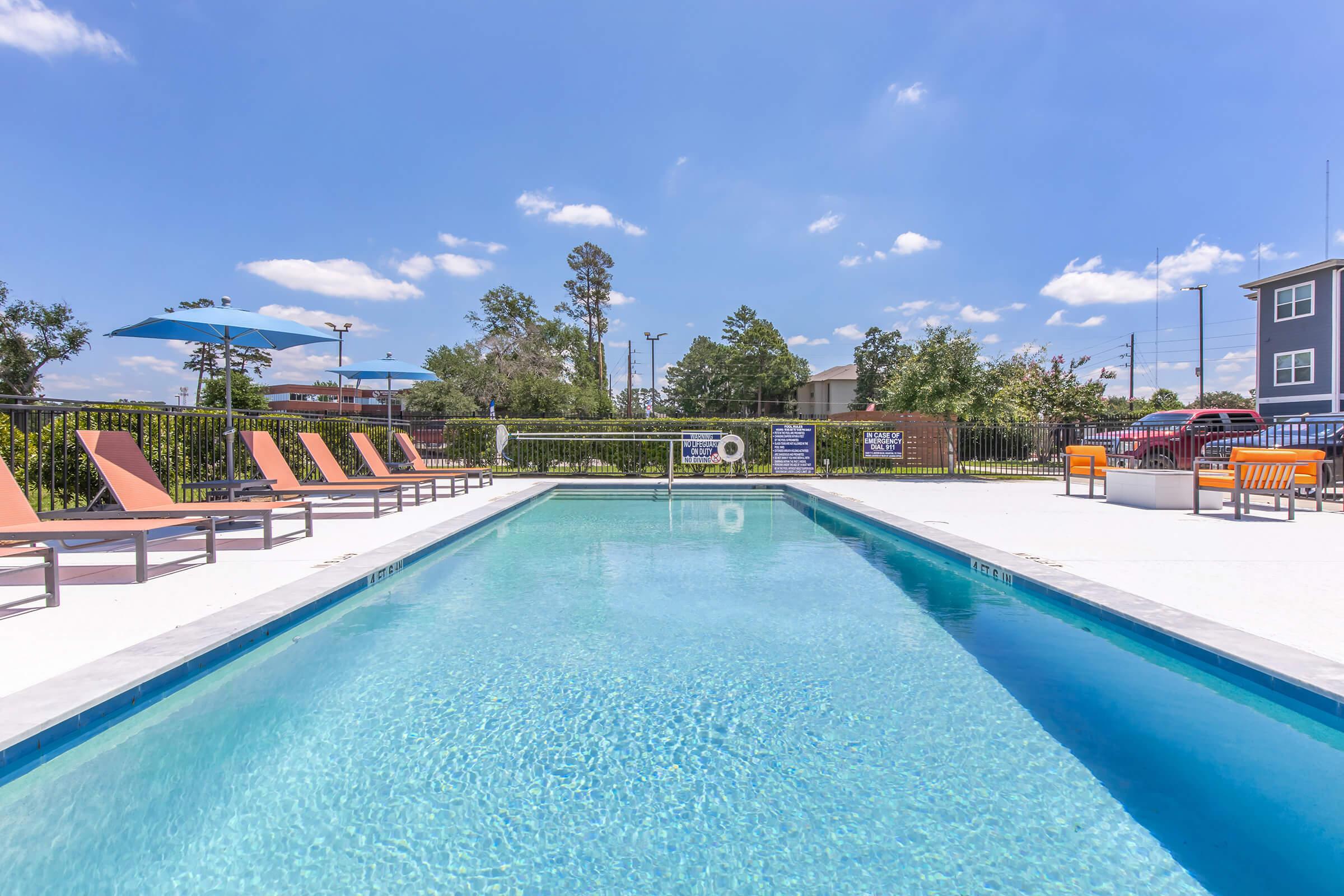 A clear swimming pool surrounded by lounge chairs, with colorful umbrellas providing shade. Lush green landscaping borders the area, and the sky is bright with a few fluffy clouds. The scene suggests a relaxing outdoor environment suitable for leisure activities.