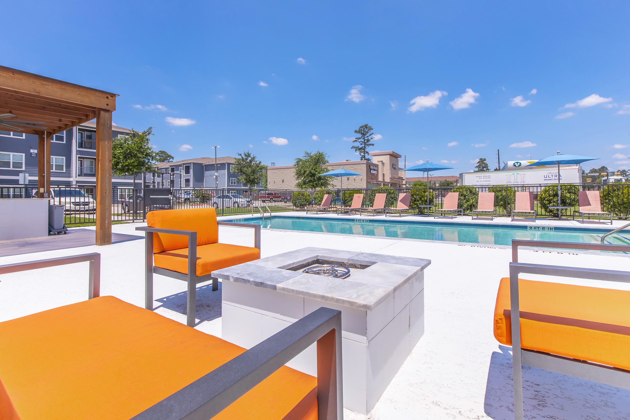 A bright and sunny poolside area featuring orange outdoor furniture, including chairs around a fire pit. In the background, there are lounge chairs by the pool and several tall trees and buildings. The clear blue sky adds to the relaxing atmosphere of the space.