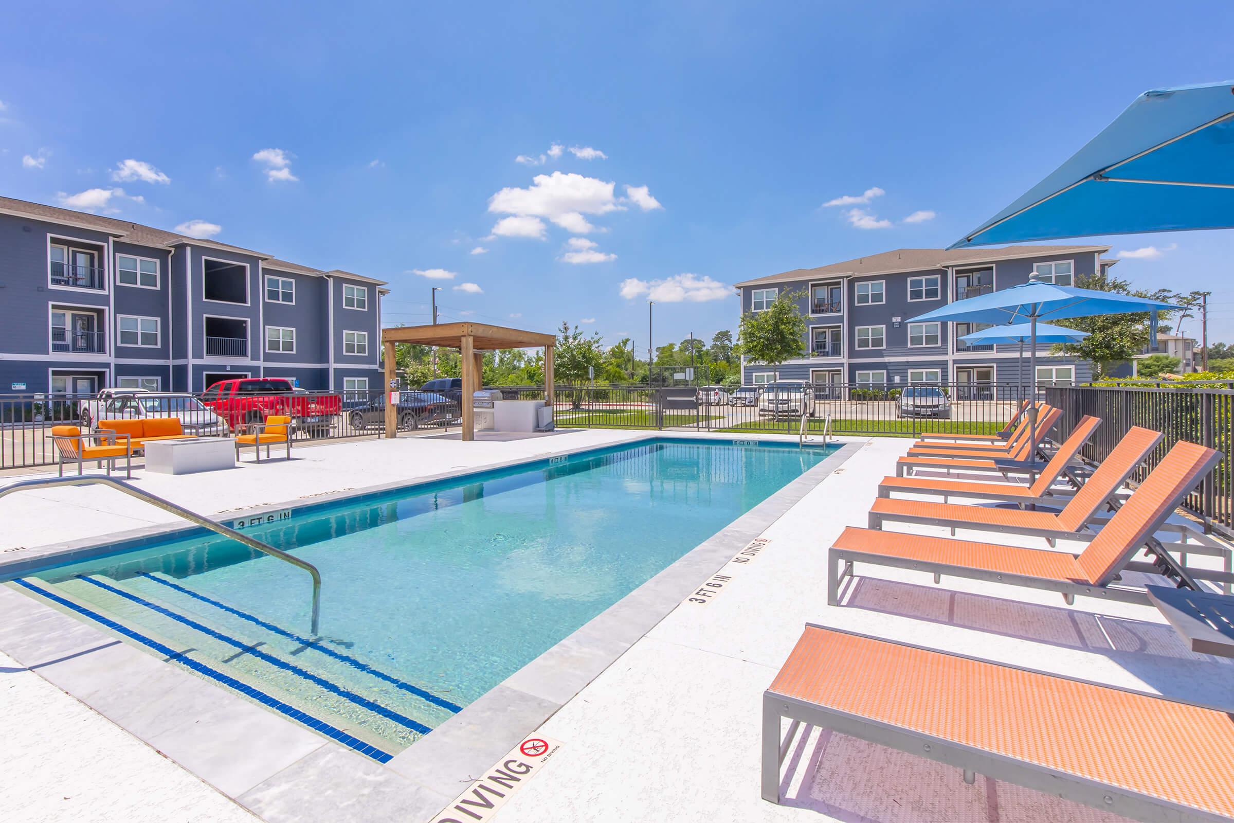 A clear blue swimming pool surrounded by orange lounge chairs and umbrellas, with an apartment complex visible in the background. The pool area is sunny, featuring a fenced perimeter and a shaded seating area with a pergola. The setting suggests a resort-like atmosphere.