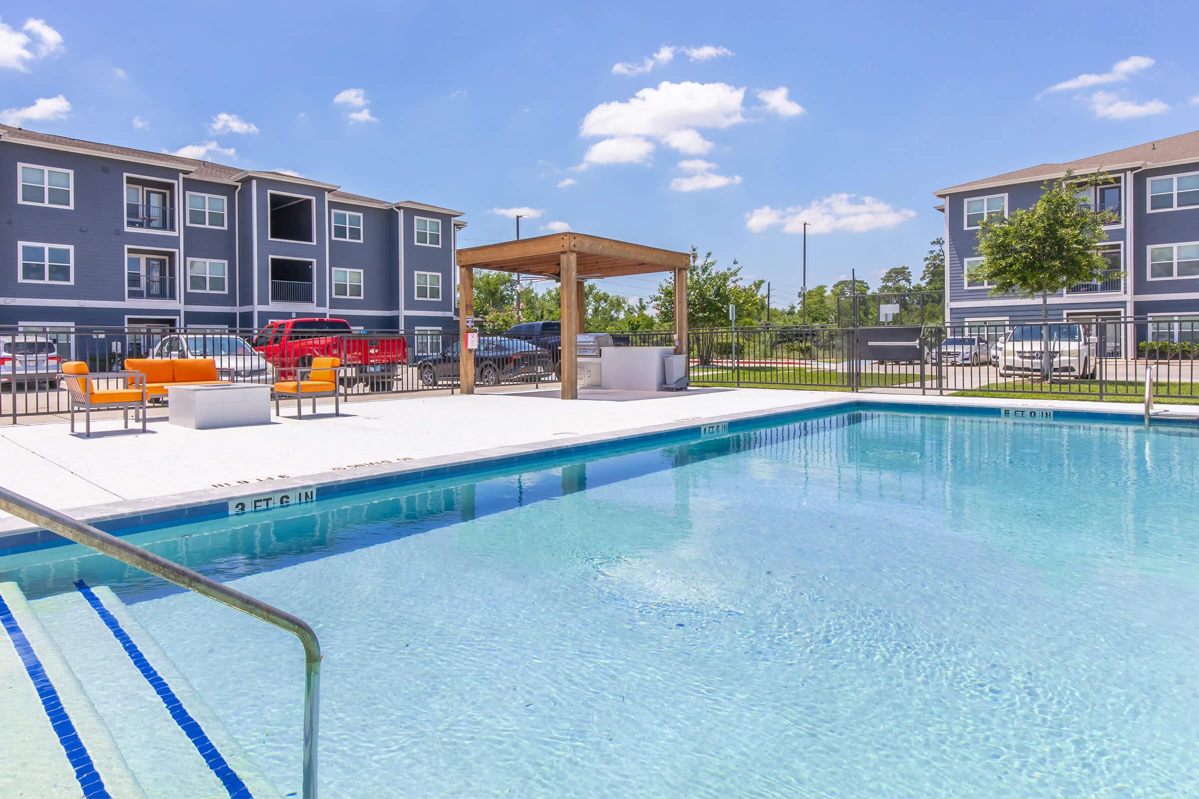 A clear blue swimming pool surrounded by a white deck, featuring orange lounge chairs and a shaded seating area with a pergola. In the background, modern apartment buildings and a green grassy area are visible under a bright blue sky with fluffy clouds.