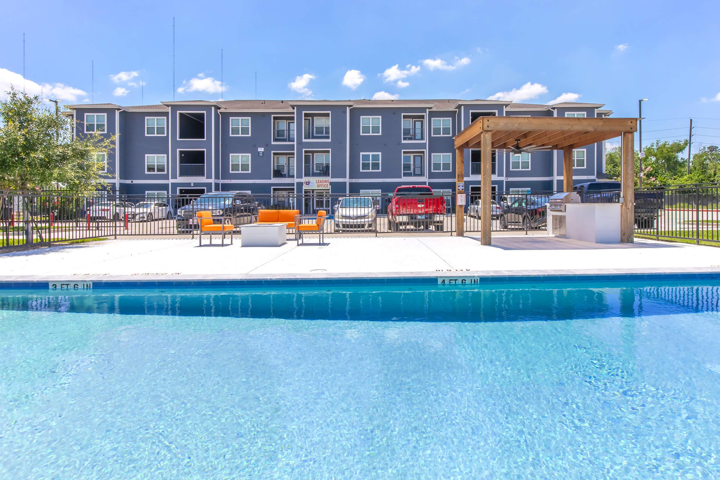 A clear blue swimming pool with a shallow area in the foreground, surrounded by a white deck. In the background, a modern apartment building with multiple balconies is visible. A pergola with seating is located near the pool, and cars are parked nearby under a bright blue sky with a few clouds.