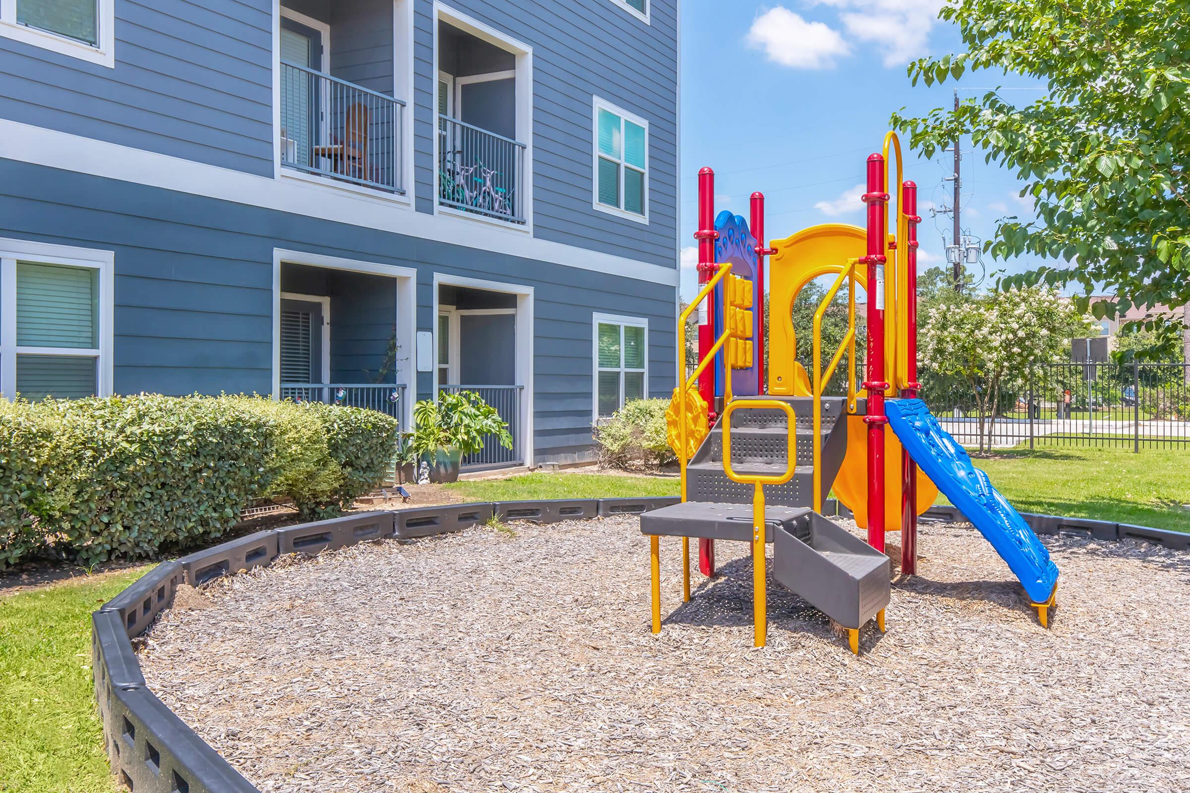 A colorful playground featuring a slide and climbing structure surrounded by mulch, set against a backdrop of a multi-story apartment building. Green shrubs and trees provide a natural border around the play area, with a clear blue sky above.