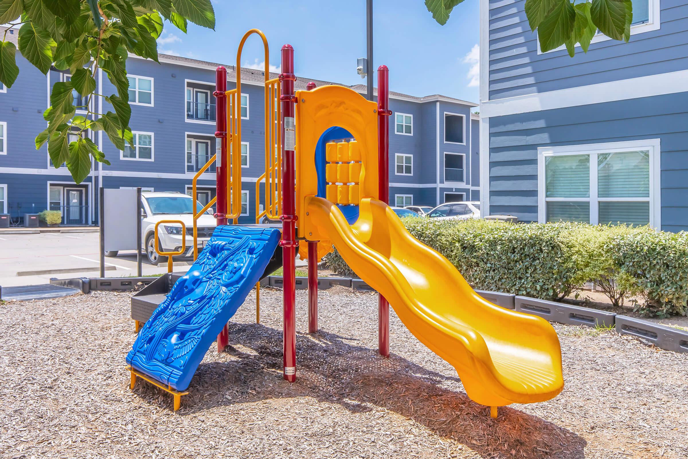 A colorful playground structure featuring a blue slide and an orange climbing frame with a ladder. The playground is surrounded by mulch and located near modern apartment buildings. Green leaves from trees provide shade in the foreground. Bright blue sky with a few clouds.