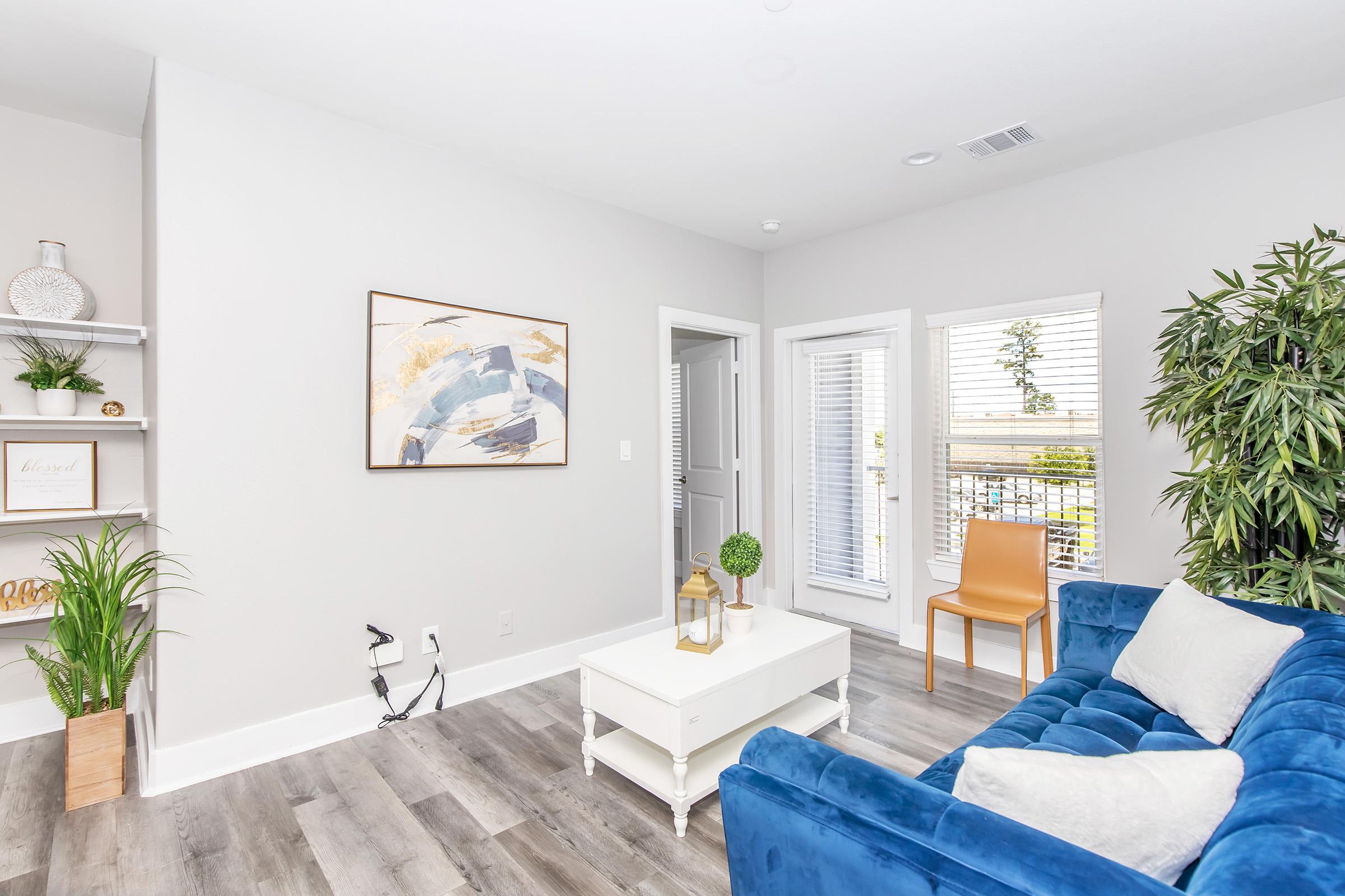 A modern living room featuring a blue velvet sofa, a white coffee table, and a decorative plant. There is a wall-mounted shelf with framed art and greenery. Natural light streams in through large windows with white blinds, and a tan chair is positioned nearby, adding to the cozy atmosphere.