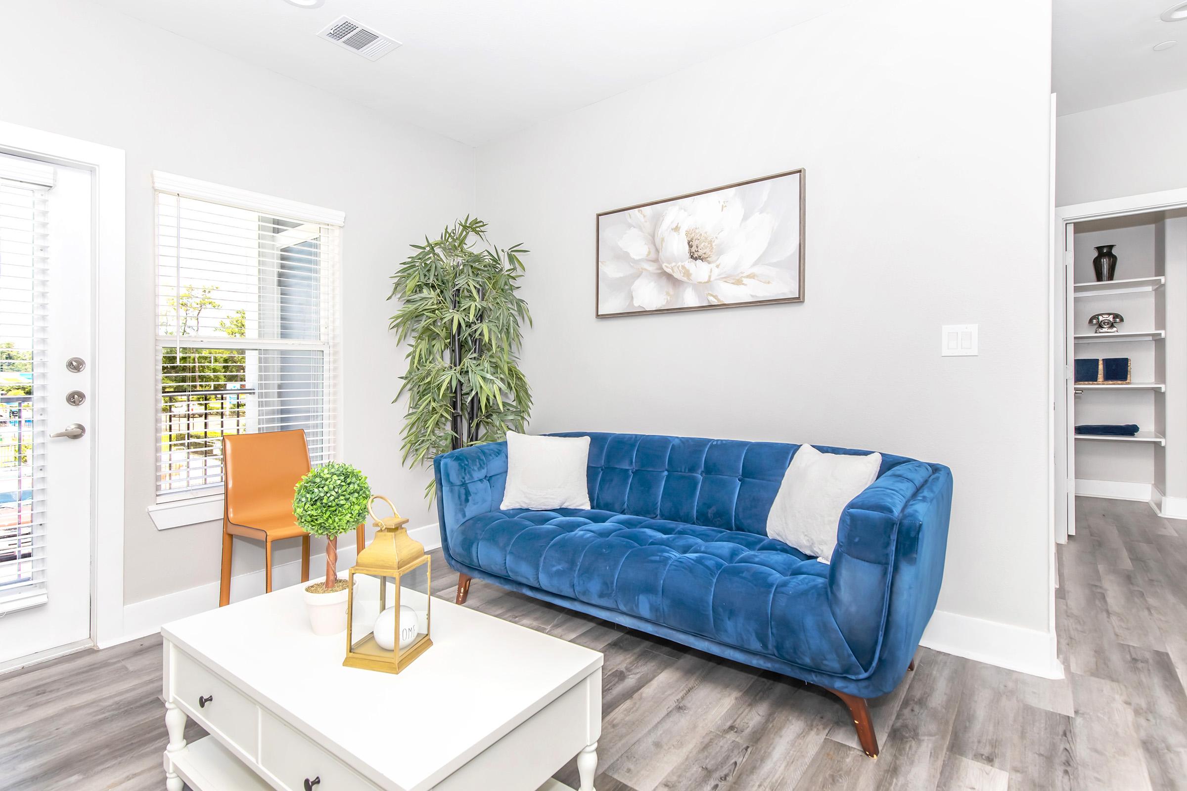 A modern living room featuring a blue velvet sofa with two white cushions, a small coffee table, and a decorative lantern. There's a potted plant in the corner, an orange chair, and a framed flower painting on the wall. Large windows allow natural light, enhancing the bright atmosphere.