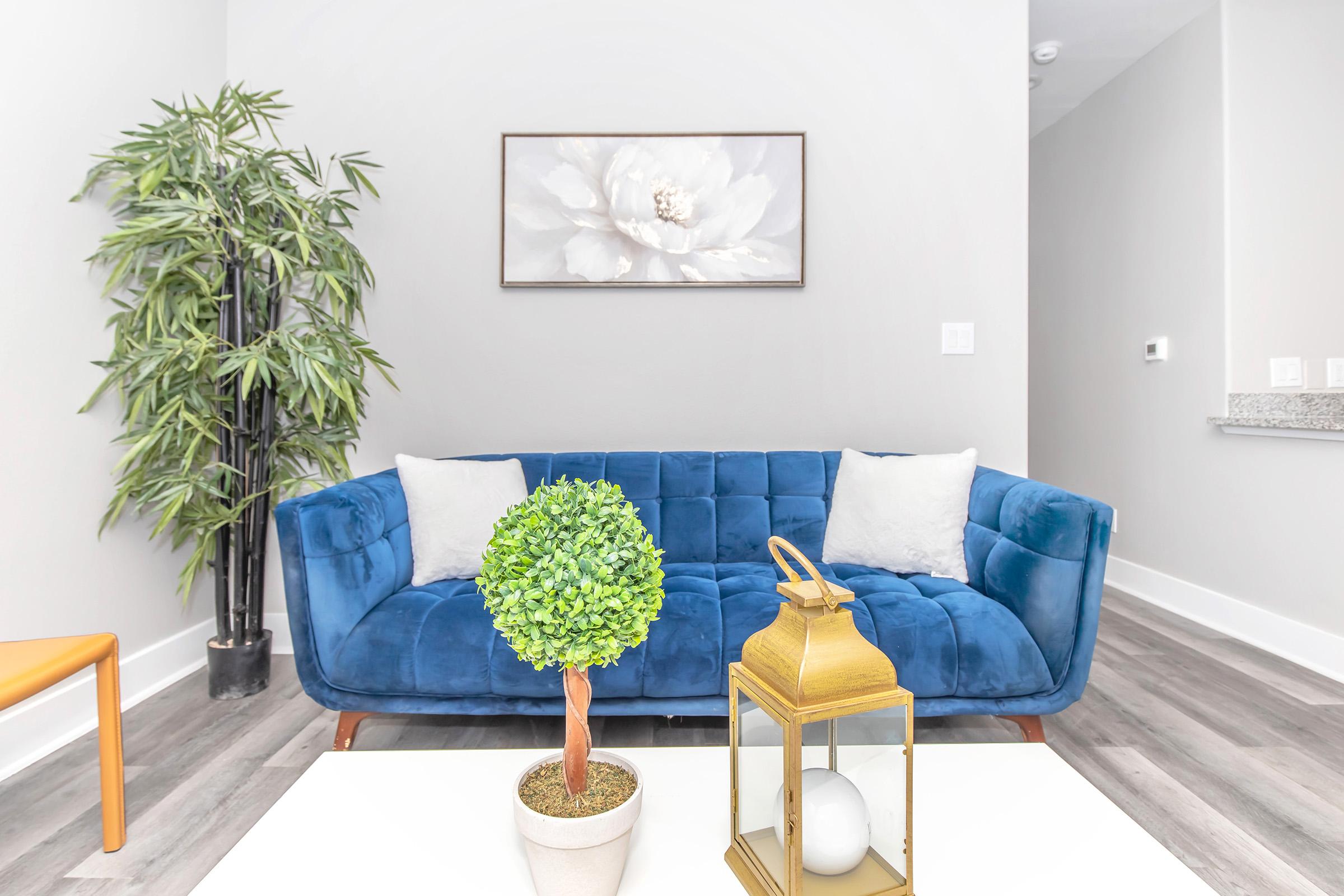 A modern living room featuring a blue velvet sofa with two light-colored cushions, a small round table with a decorative potted plant and a lantern, alongside a tall indoor plant. A wall art piece with a flower design is hung above the sofa, and the room has light gray walls and wooden flooring.