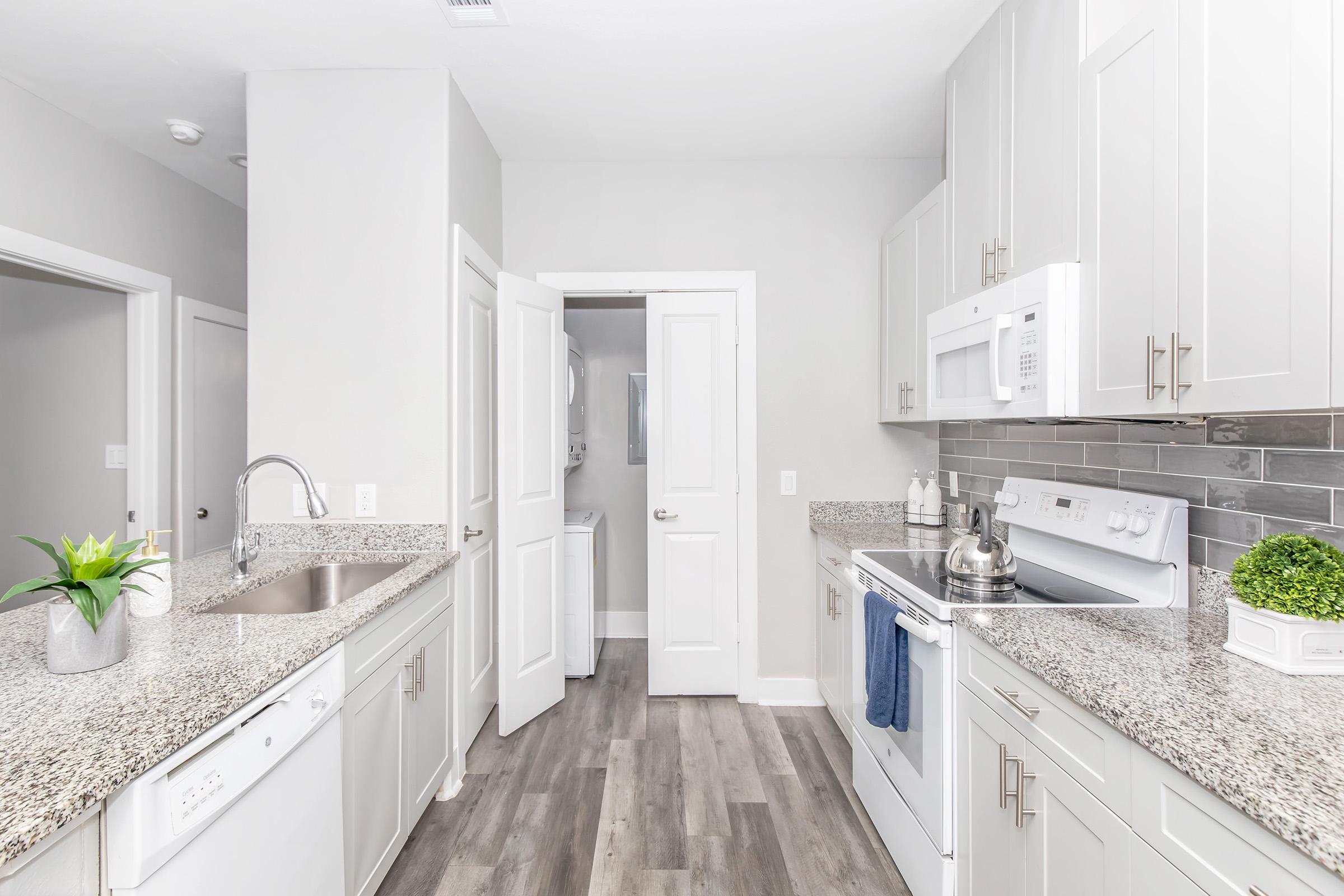 Modern kitchen featuring light gray cabinets and granite countertops, stainless steel appliances including a microwave and stove, and a neatly arranged sink with a small plant on the counter. The space has a door leading to a laundry area and a neutral color scheme, enhancing the bright and airy atmosphere.