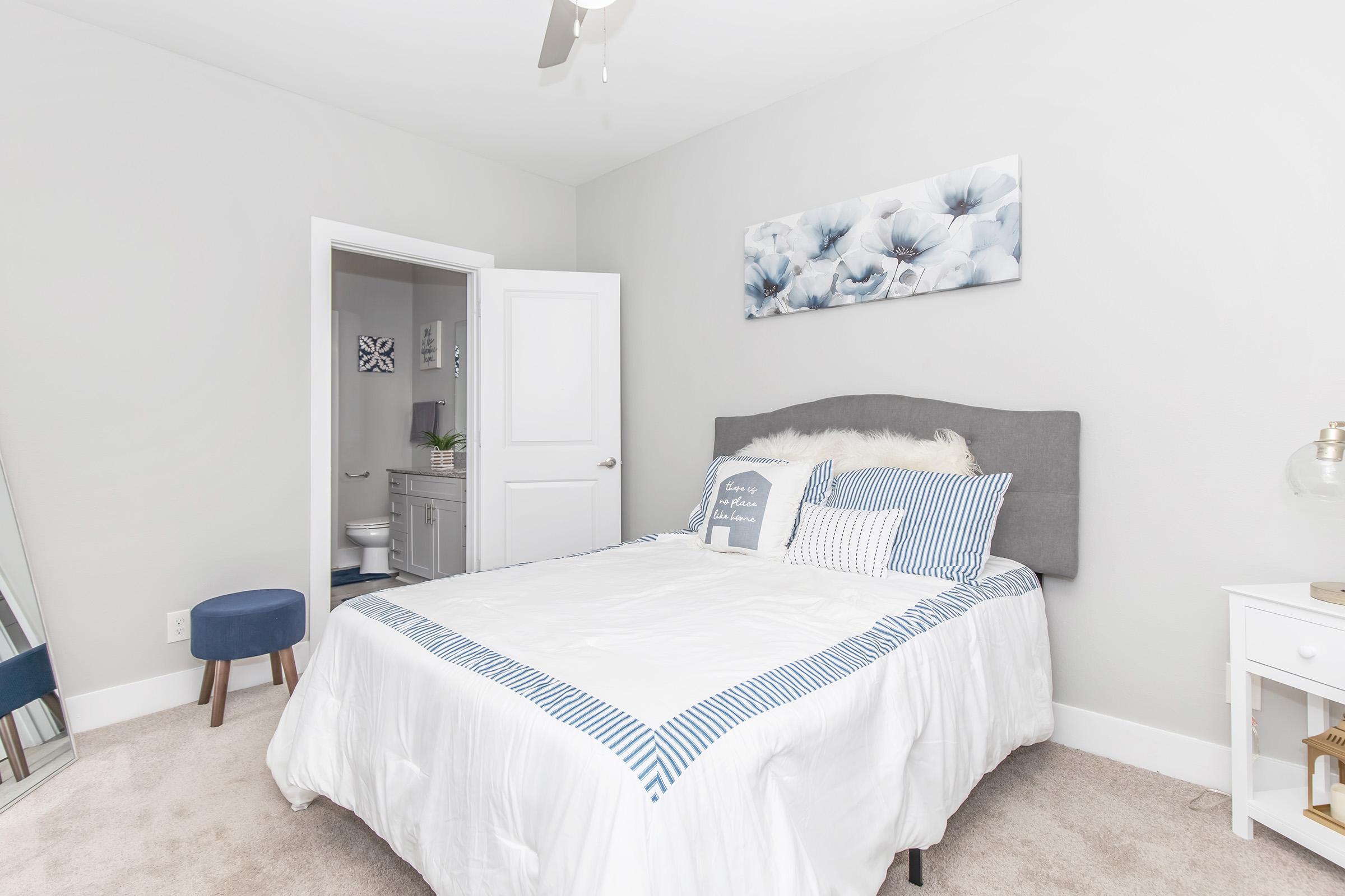A cozy, modern bedroom featuring a gray upholstered bed with blue and white striped bedding. A light gray wall with a floral painting above the bed, a small blue stool, and a white nightstand with a lamp. A bathroom door is visible in the background, and a large mirror reflects the space.