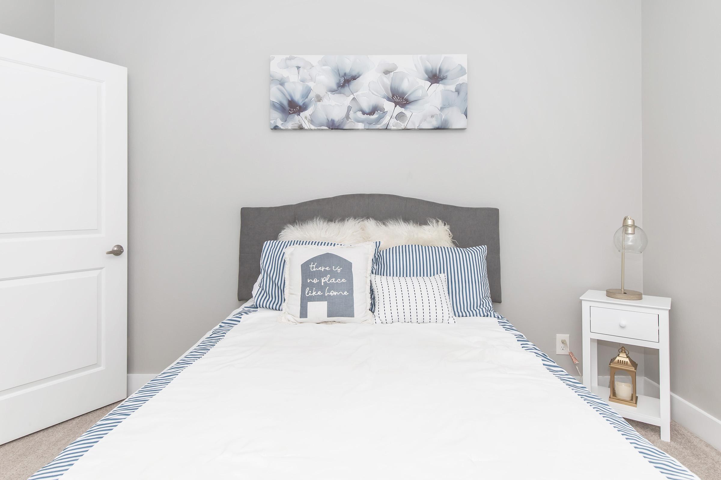 A neatly made bed with blue and white striped pillows and a decorative throw pillow featuring the phrase "there is no place like home." Above the bed is a floral wall art piece. A bedside table with a lamp and decorative item is beside the bed, set in a light-colored room with gray walls.
