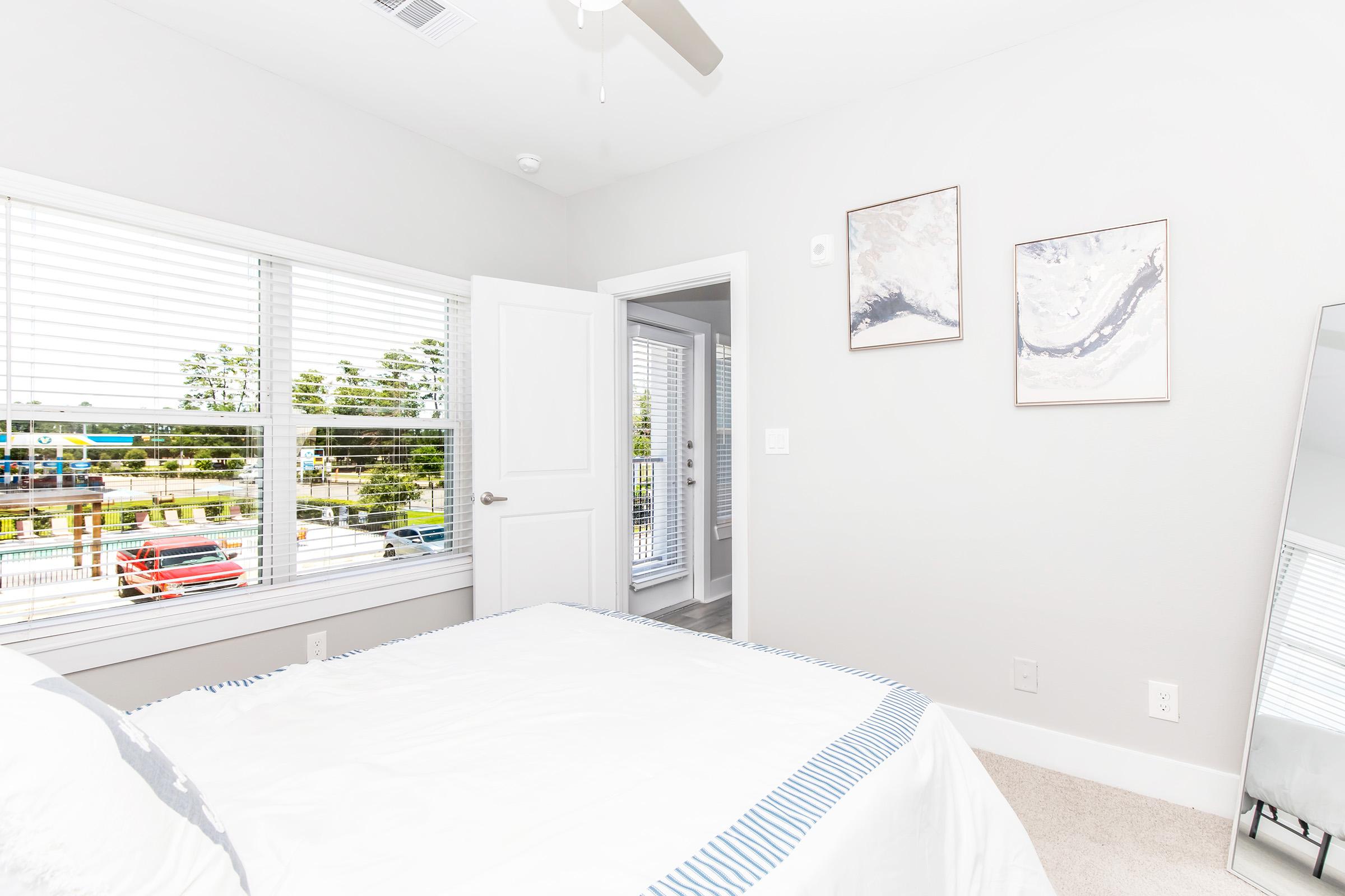 A bright bedroom featuring a white bed with a striped blanket, large window with blinds, minimalist decor, and two framed pictures on the wall. A full-length mirror stands to the side, and a view of parked cars and greenery is visible outside the window.