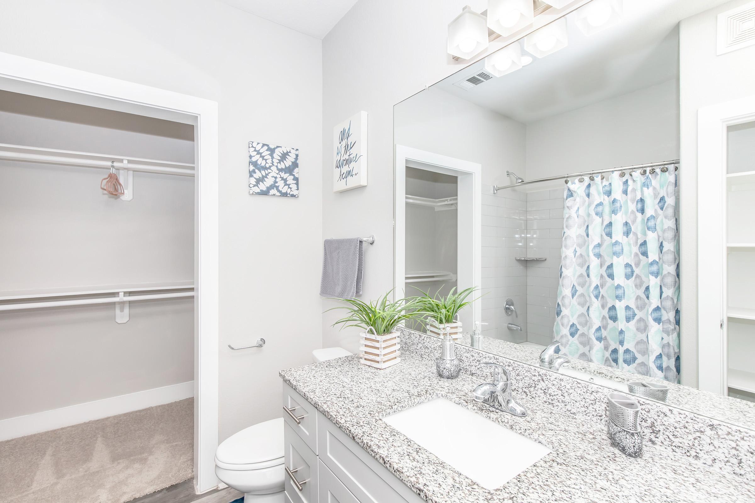 A modern bathroom featuring a large mirror above a granite countertop with a white sink. There is a shower with a patterned blue and white curtain, and shelves in the background. The space is decorated with plants and artwork, with neutral-colored walls and storage areas nearby.