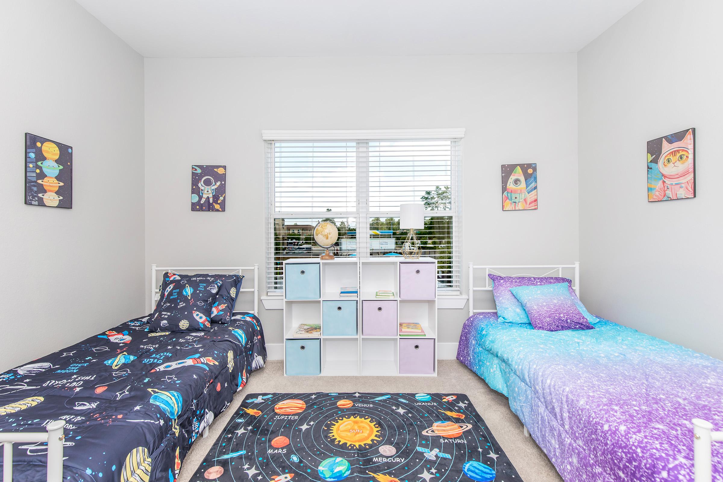 A children's bedroom featuring two twin beds with space-themed bedding, a colorful area rug with a planetary design, and wall art of planets and rockets. A white bookshelf holds various items and is placed under a window with natural light. The room has a light gray color scheme.