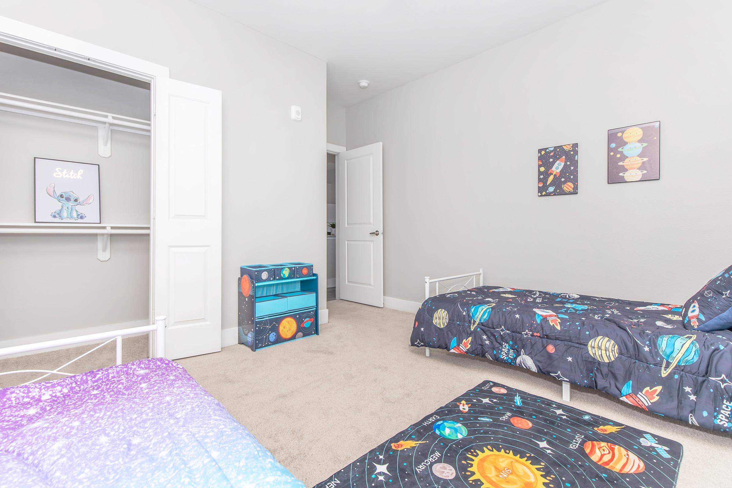 A children's bedroom featuring two twin beds with space-themed bedding. One bed is purple, and the other is blue, both decorated with planets and stars. There is a small blue dresser, a closet with sliding doors, and two framed space-themed artworks on the wall. The room has a light gray color scheme.