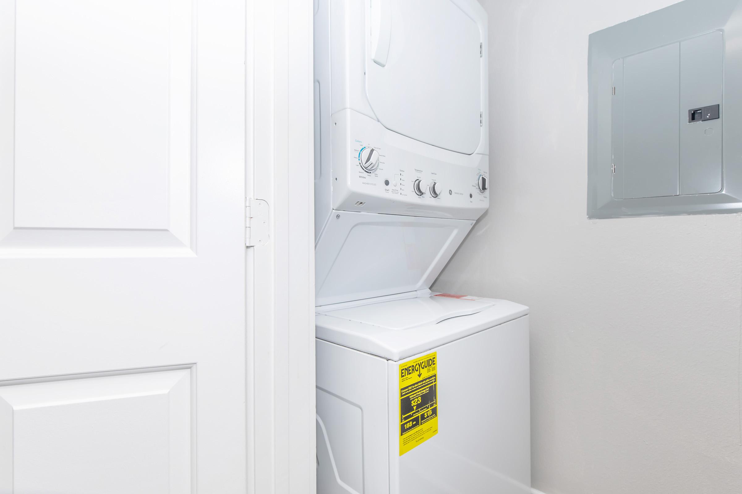 A stacked washer and dryer unit in a small laundry space next to a white door. A warning label is visible on the dryer. The wall is painted light gray, and there is an electrical panel nearby. The overall area appears clean and organized.