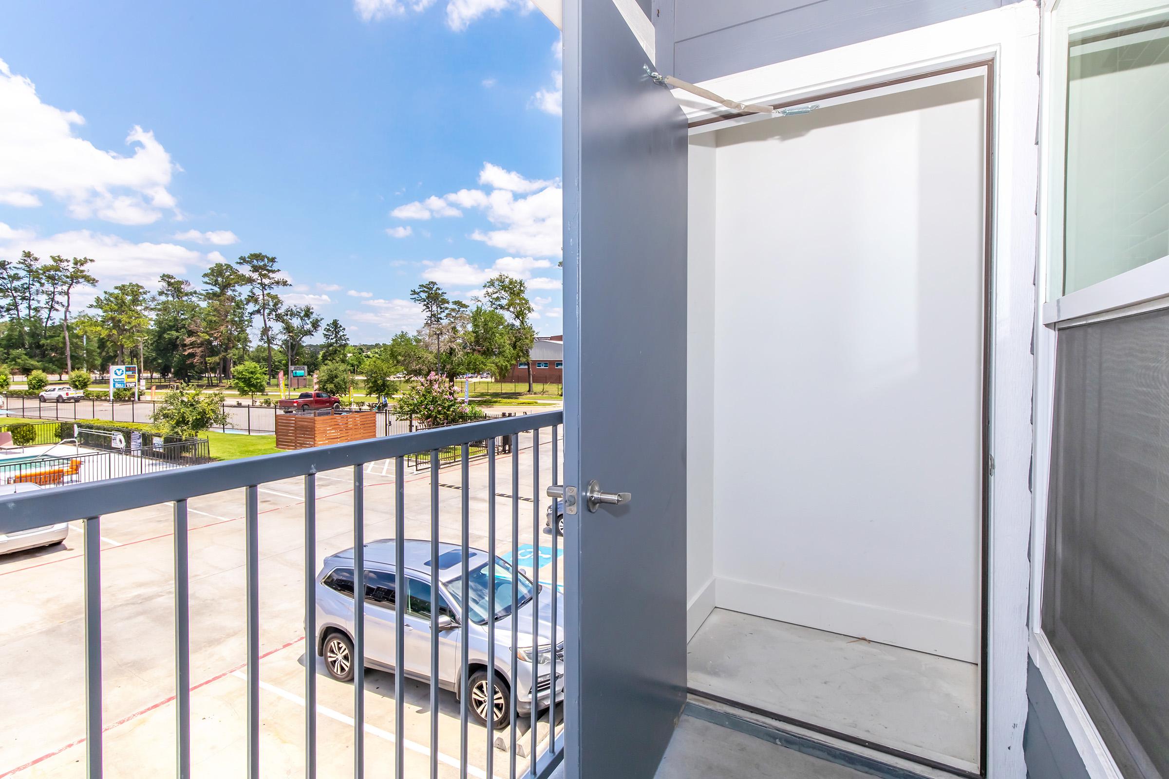 A view from a balcony looking at an open door leading to a storage closet. The balcony features a metal railing and overlooks a parking area with a gray car parked below. Surrounding trees and a bright blue sky with fluffy clouds are visible in the background.
